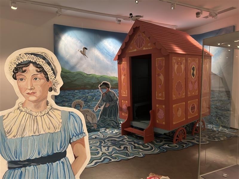 Museum display featuring wooden bathing machine hut with dark interior, life-sized cutout of woman in blue dress and white bonnet.