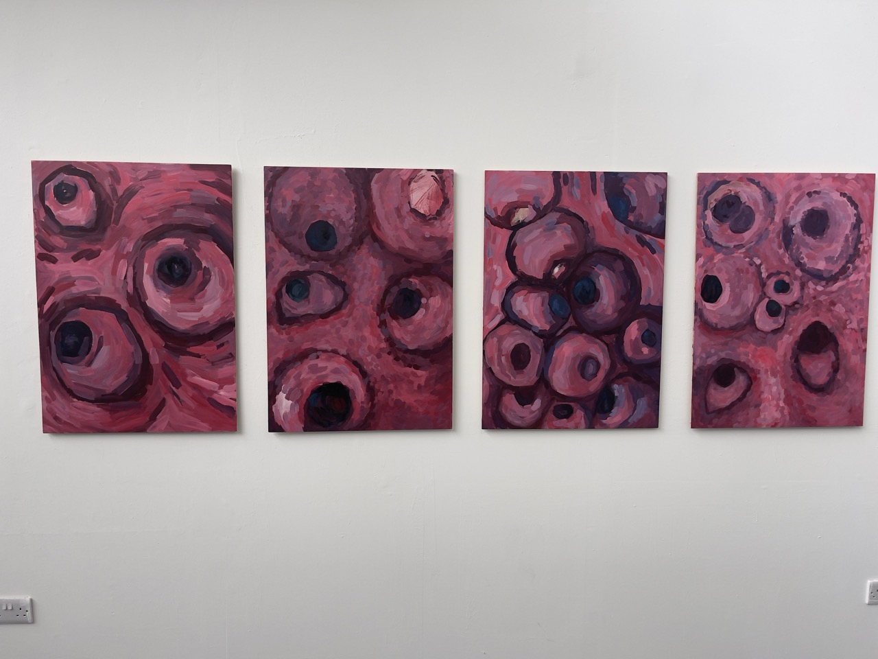 Four abstract paintings with a predominant pink and red colour palette, featuring circular shapes and patterns. The artworks are displayed side by side on a white wall.