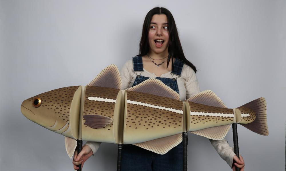 AUB student looking surprised holding up a model of a huge brown-yellow cod with two handles underneath, separated into four separate sections.