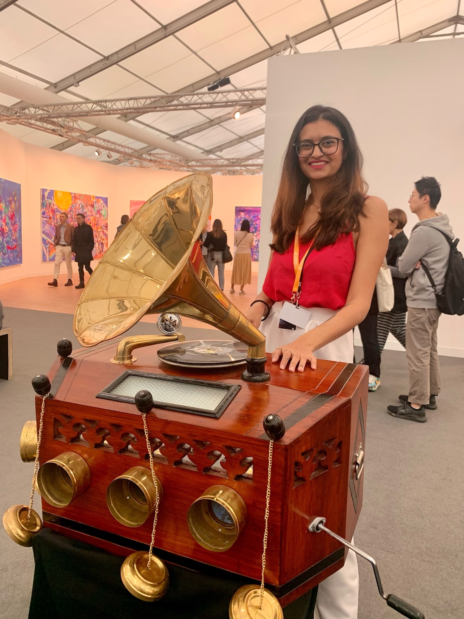 Woman in red top standing behind ornate wooden gramophone with brass horn and dials at art gallery exhibition.