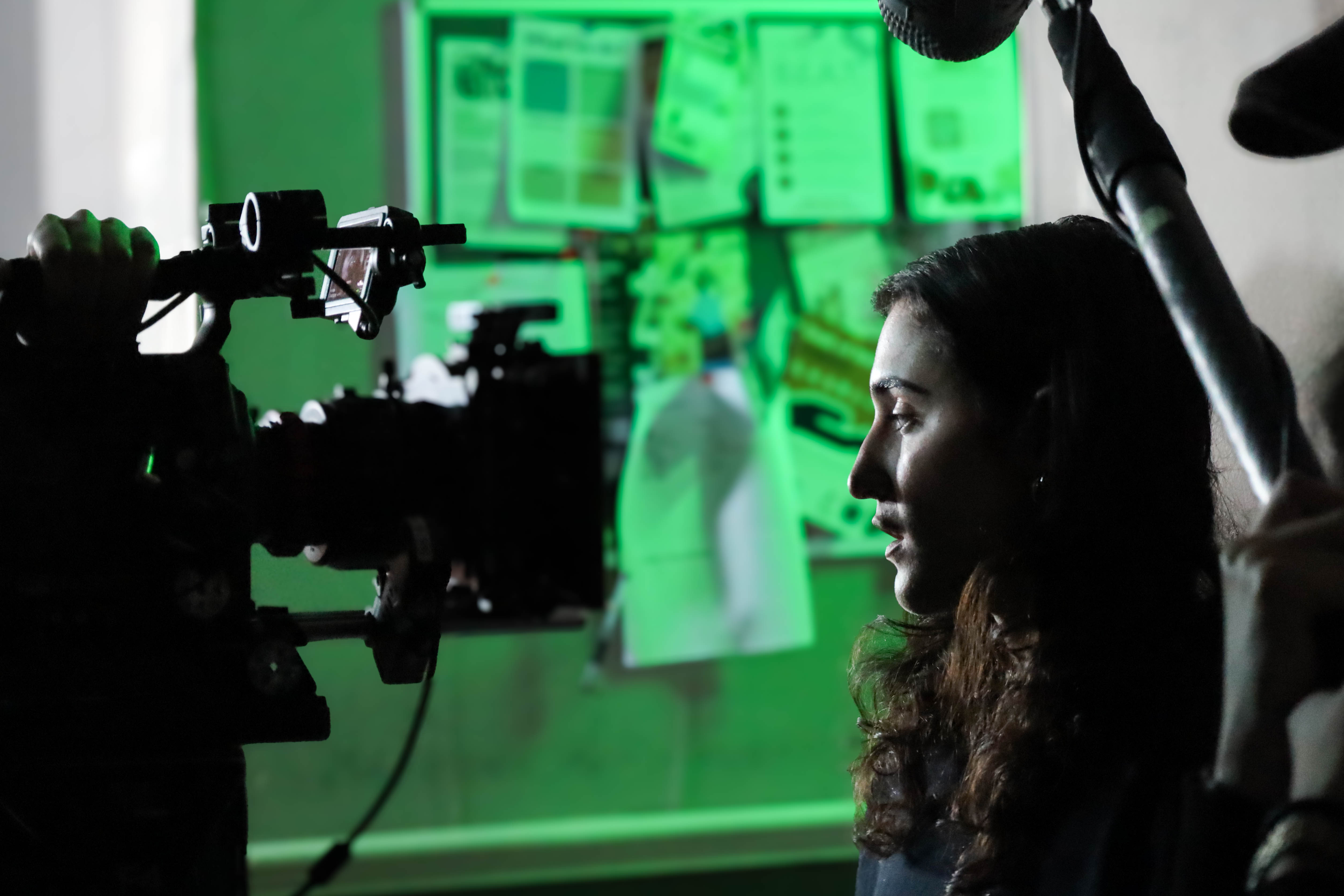 A woman with long, dark hair is positioned in front of a camera on a tripod, with a microphone above. The background features a bulletin board with various papers and printed materials, illuminated by a green light.