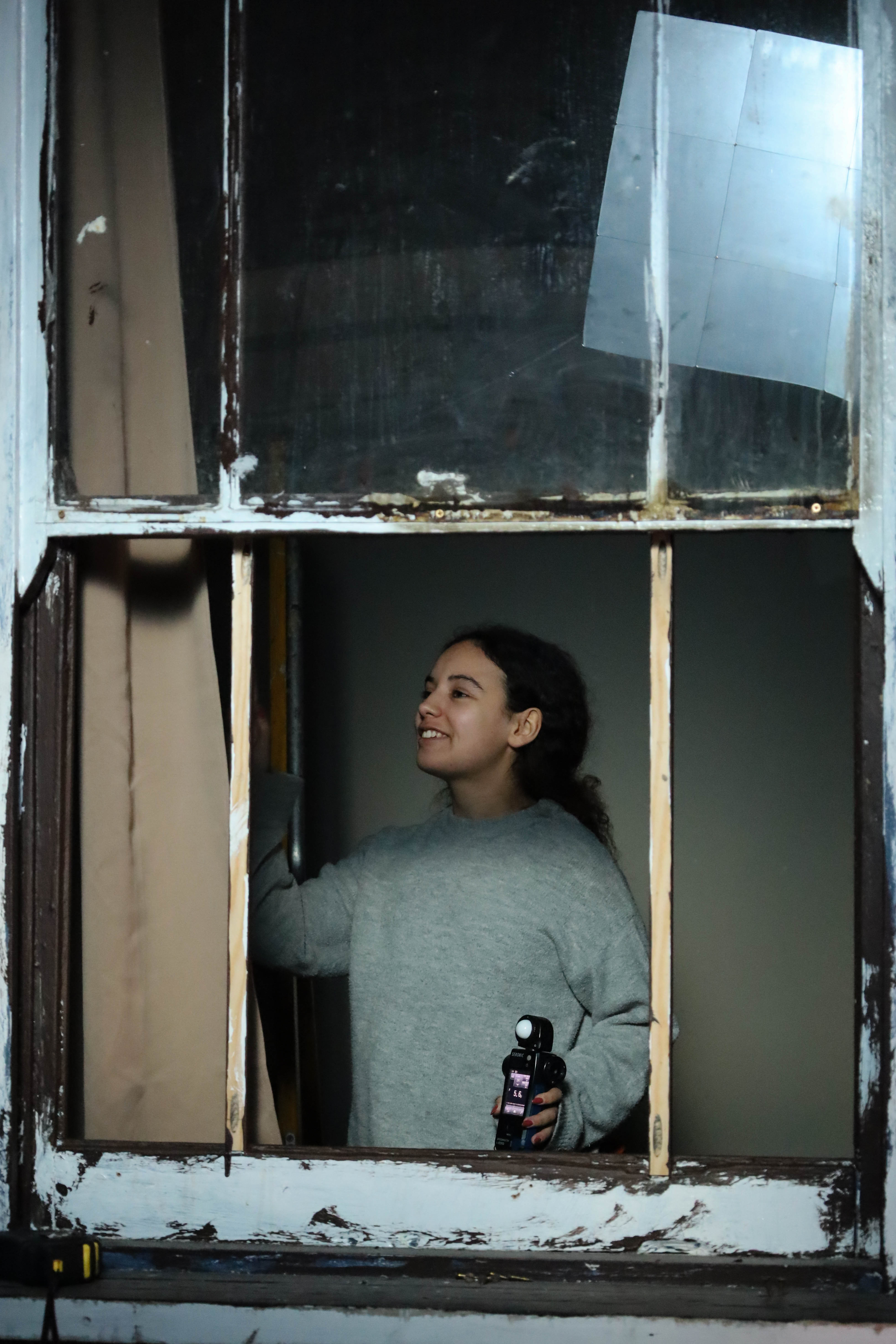 A woman wearing a grey sweater is standing by a window with peeling paint. She is pulling back a curtain and holding a bottle in her right hand, with part of her face visible through the window. The background shows a dimly lit room.