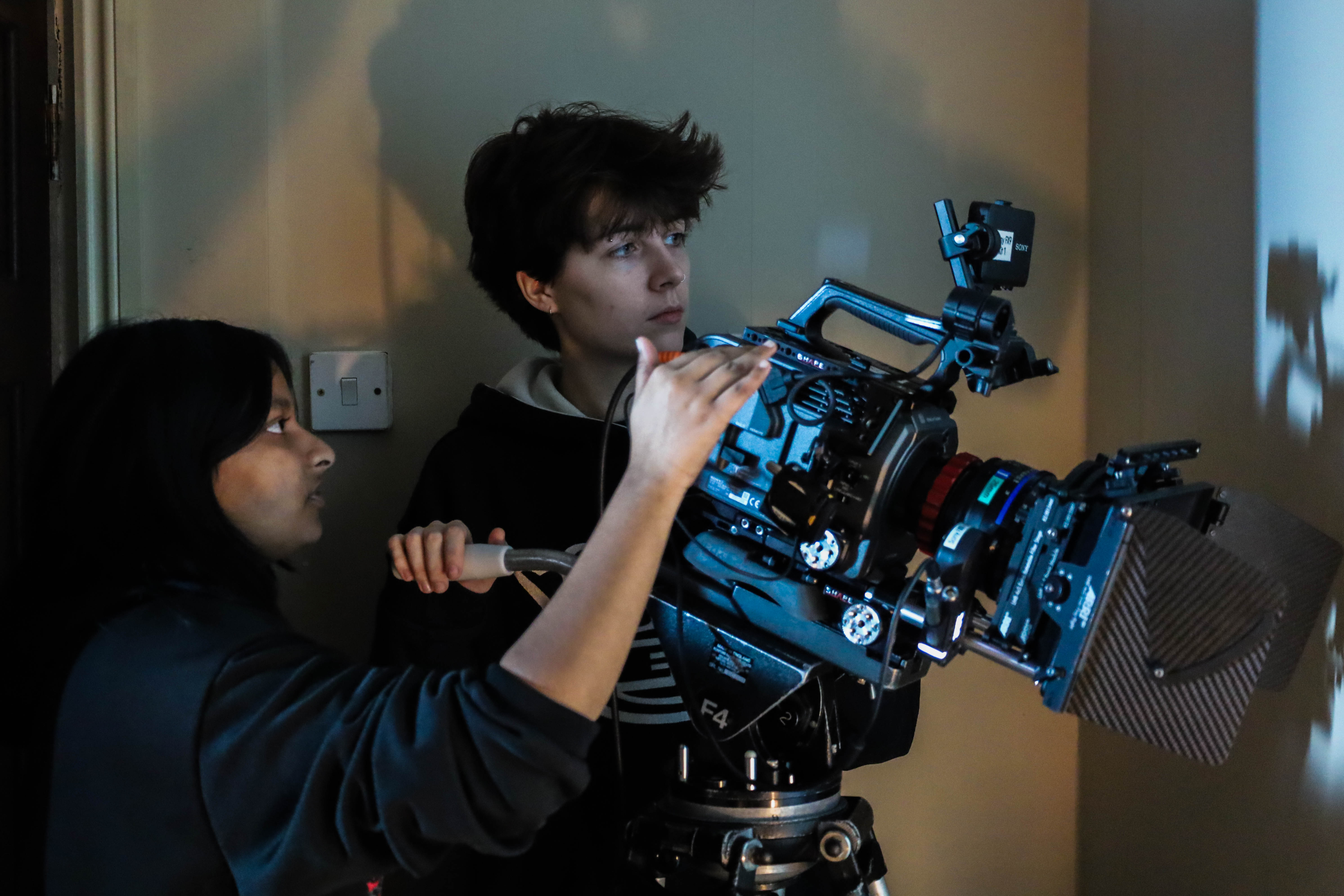 A young woman adjusts a film camera while a young man looks on attentively. The setting appears to be a room with a wall partially illuminated by projected light from the camera.