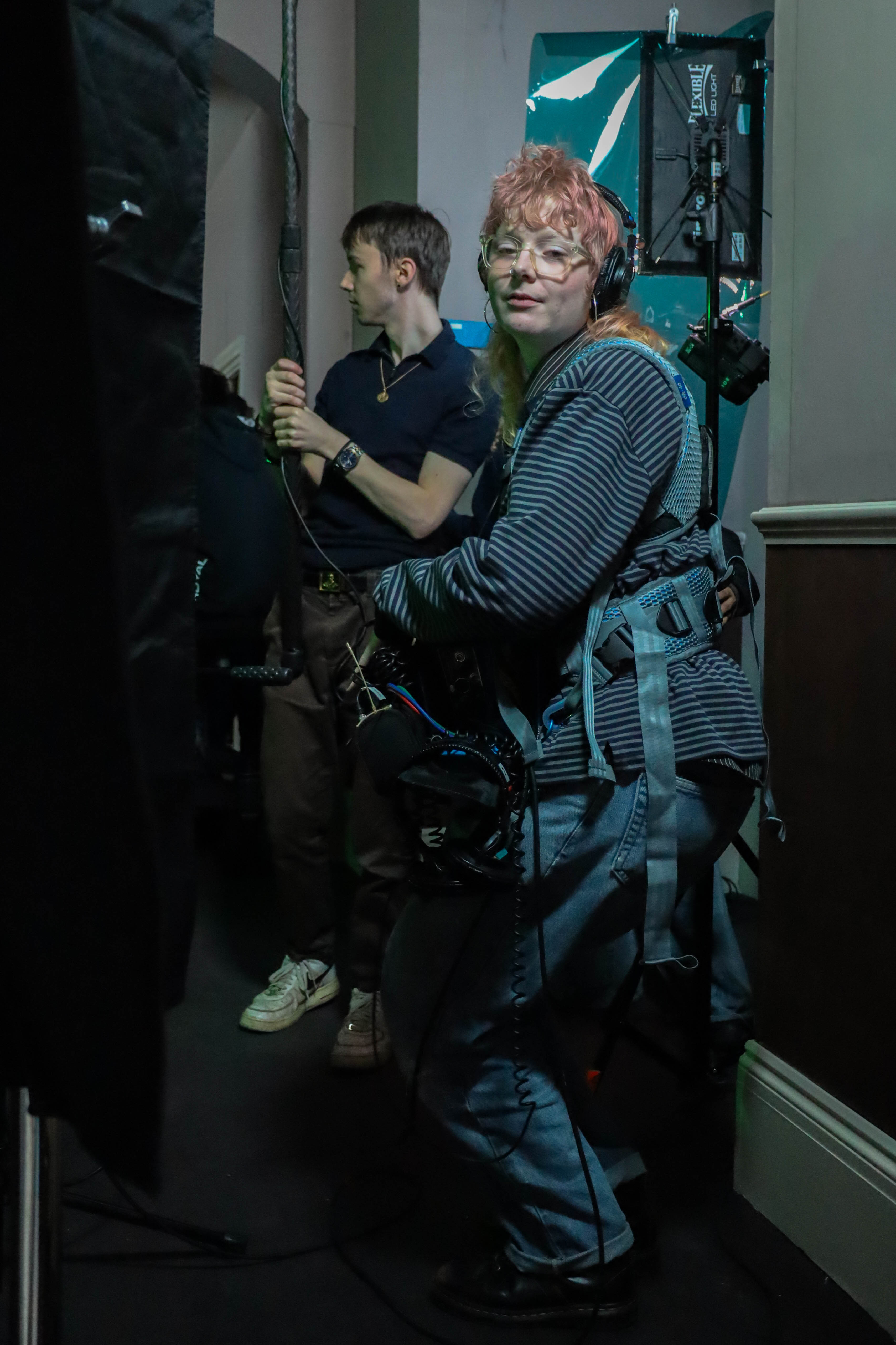 A woman with short pink hair and glasses holds filming equipment, wearing a rig and a striped sweater. In the background, a man in a navy polo shirt adjusts equipment in a dimly lit environment with a green backdrop.