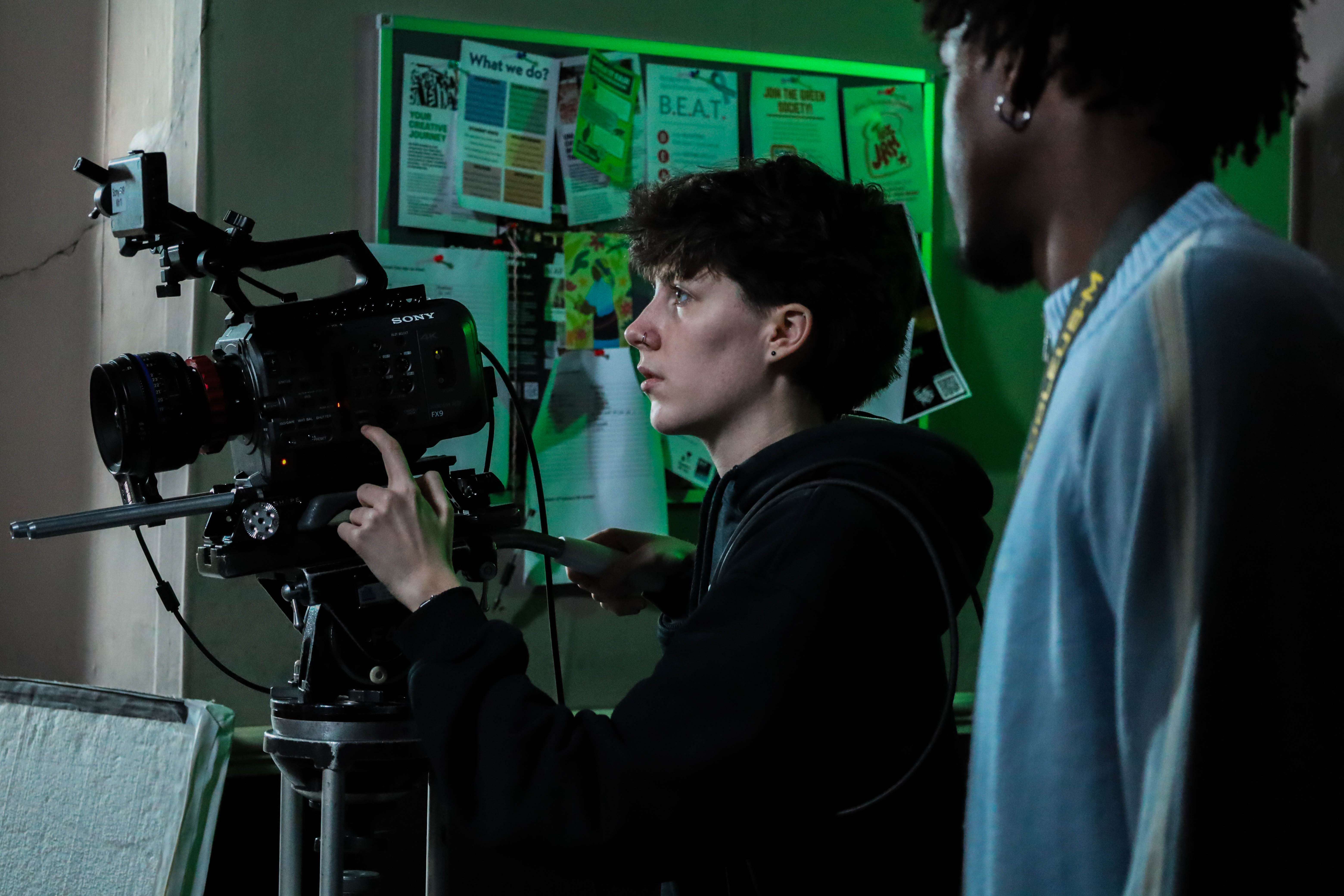 A person with short hair, dressed in a black hoodie, adjusts a camera on a tripod, focusing intently. Another person with curly hair, wearing a light blue sweater, observes from the side. The background features pinned notes and green lighting.