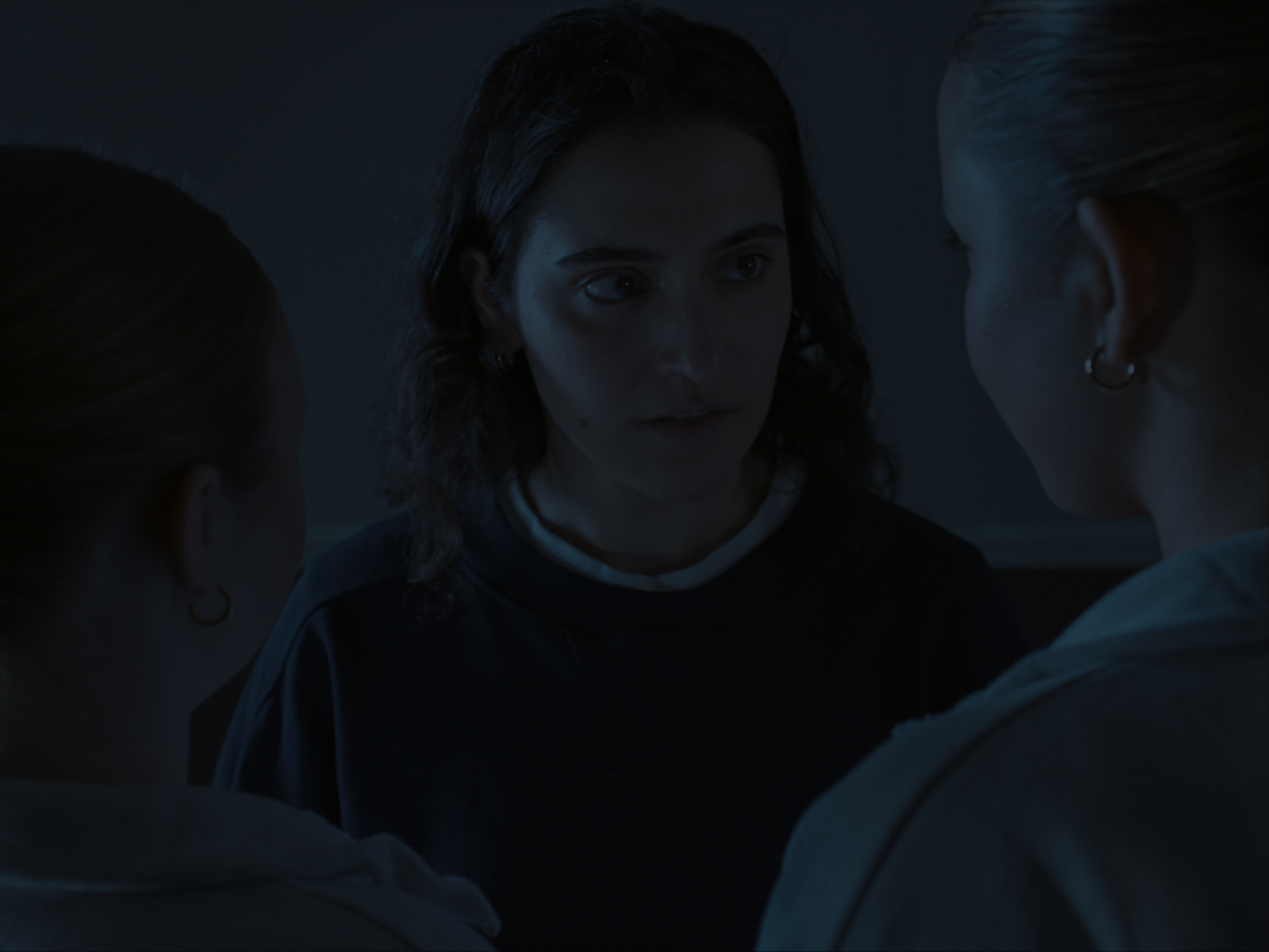 A woman with dark hair wearing a dark sweatshirt stands in a dimly lit space, facing two women. One woman on her left has a light brown bun and wears a grey top, while the other on her right has blonde hair tied back and wears a white sweatshirt.