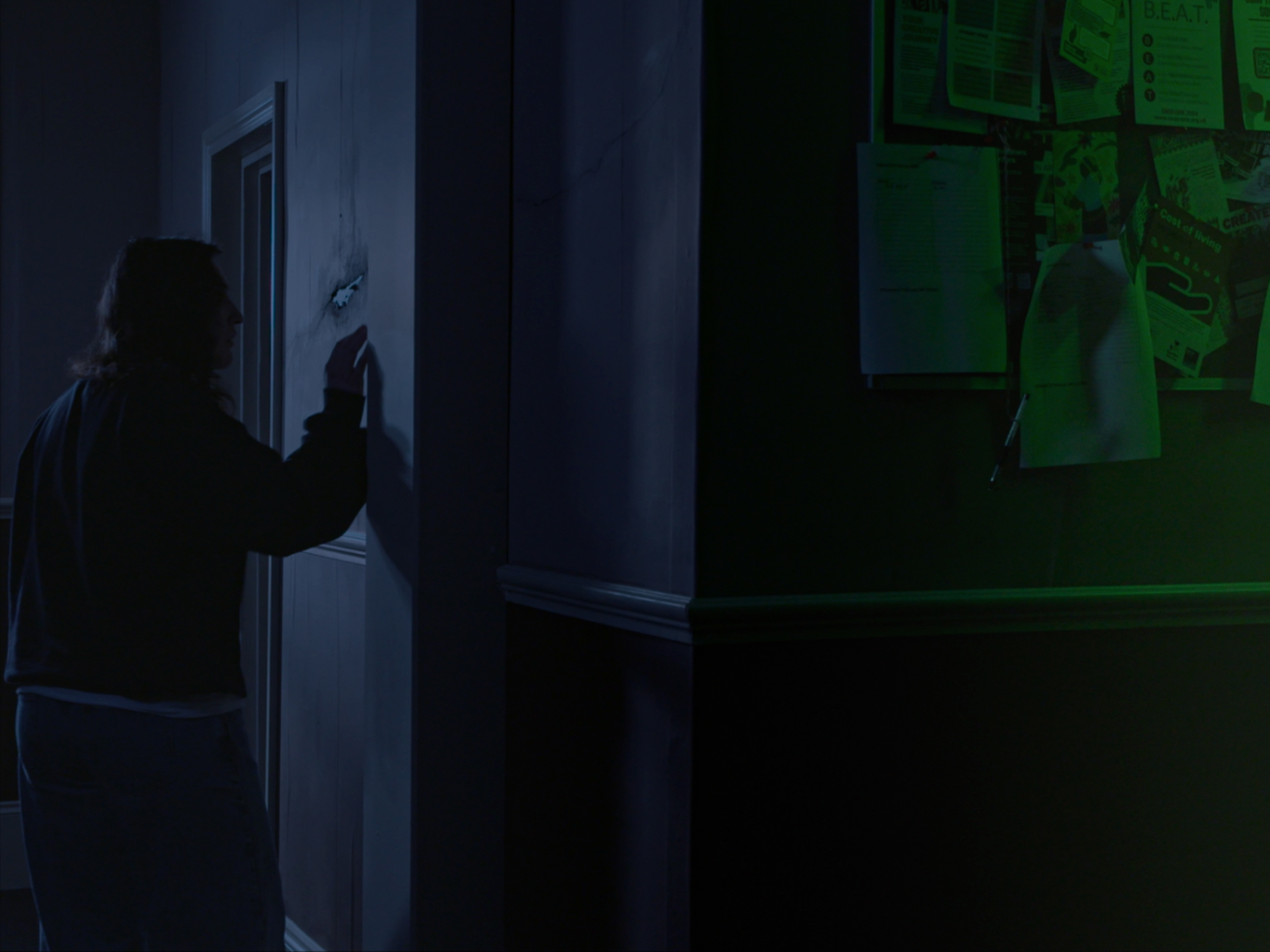 A person with long hair, dressed in a black sweater and light trousers, stands with their back to the viewer, writing on a wall in a dimly lit room. On the right, a bulletin board displays various documents and notes.