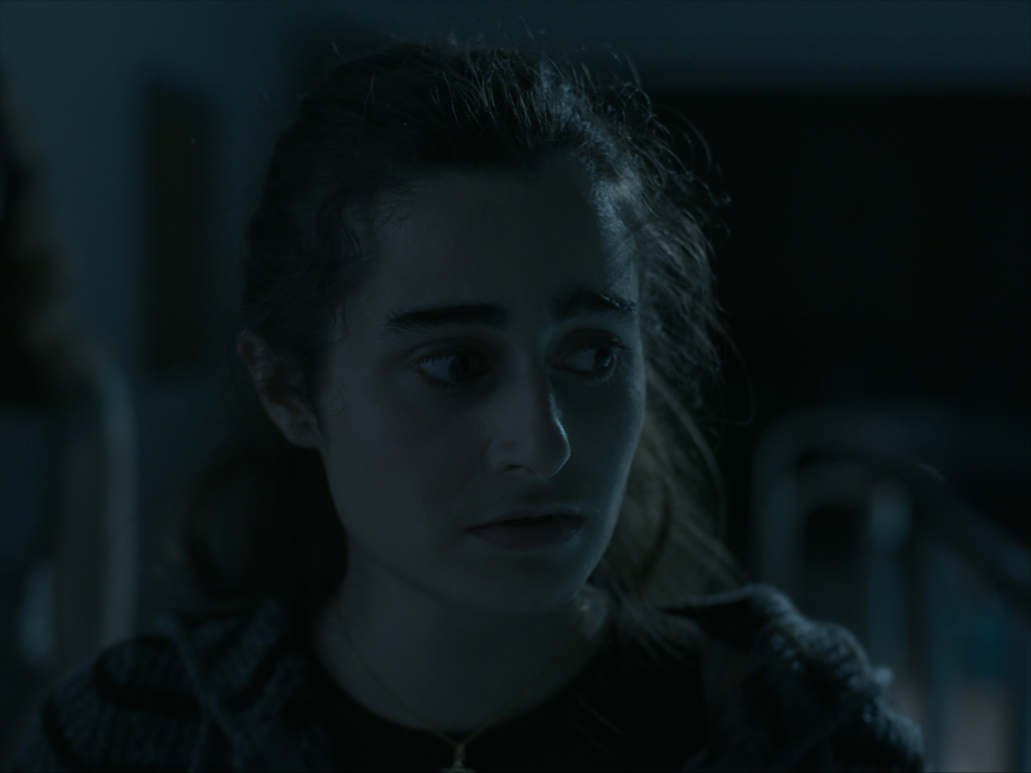 A young woman with dark hair and wearing a dark top is sitting in a dimly lit space, with a concerned expression as she looks away from the camera. The background features blurred outlines of furniture, suggesting an indoor setting.