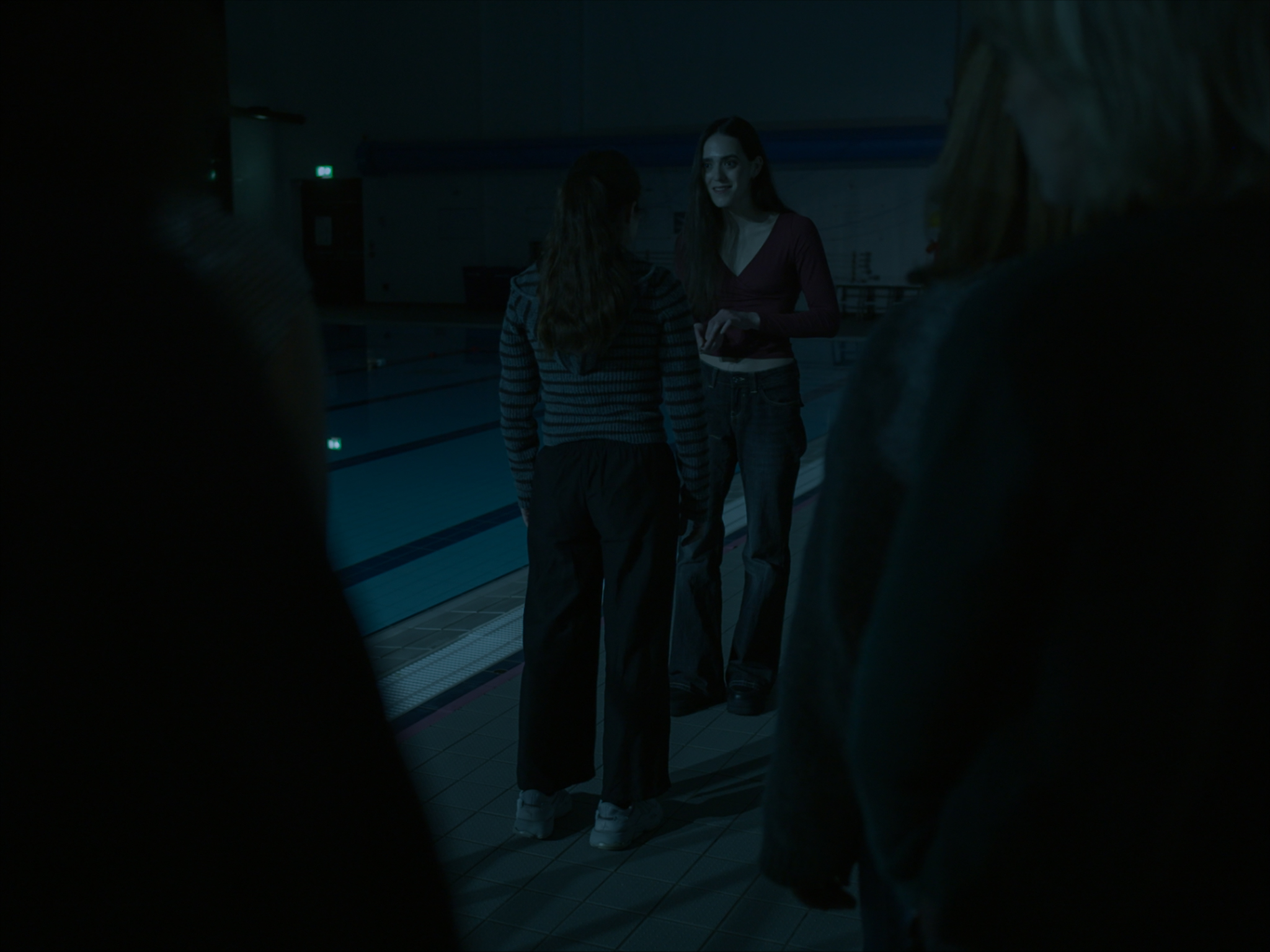 A dimly lit indoor swimming pool area where a girl in a striped top stands facing another girl in a red top. The background features other individuals partially visible, waiting beside the pool.