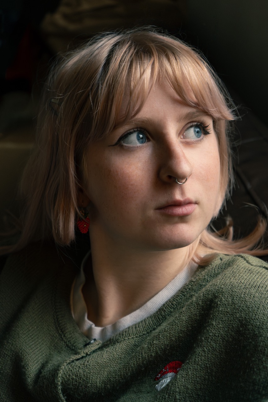 A young woman with blonde hair, styled with loose waves, looks away from the camera. She wears a green sweater and has a septum piercing along with red earrings. Soft light illuminates her face, creating gentle shadows.