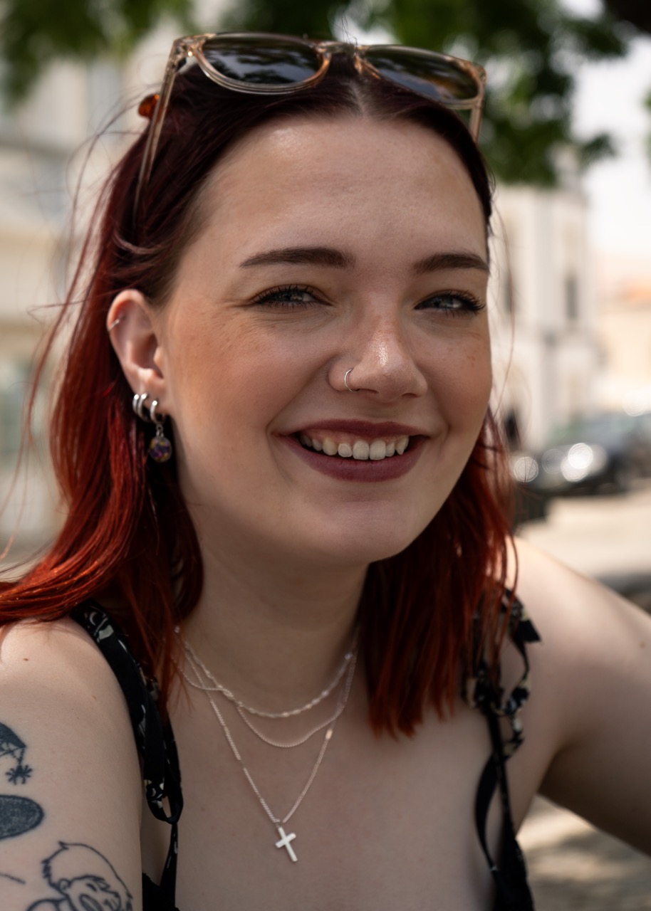 A young woman with red hair and sunglasses sits outdoors, wearing a sleeveless black top. She has multiple ear piercings and layered necklaces, with what appears to be a cross pendant. The background shows blurred cars and greenery.