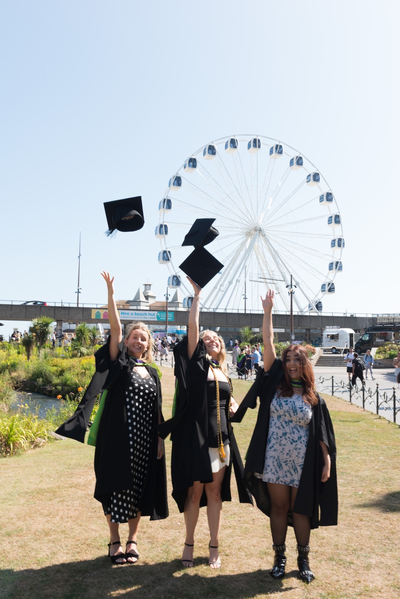 Three women in graduation gowns throwing mortarboards in air, standing on grass with large white Ferris wheel in background.