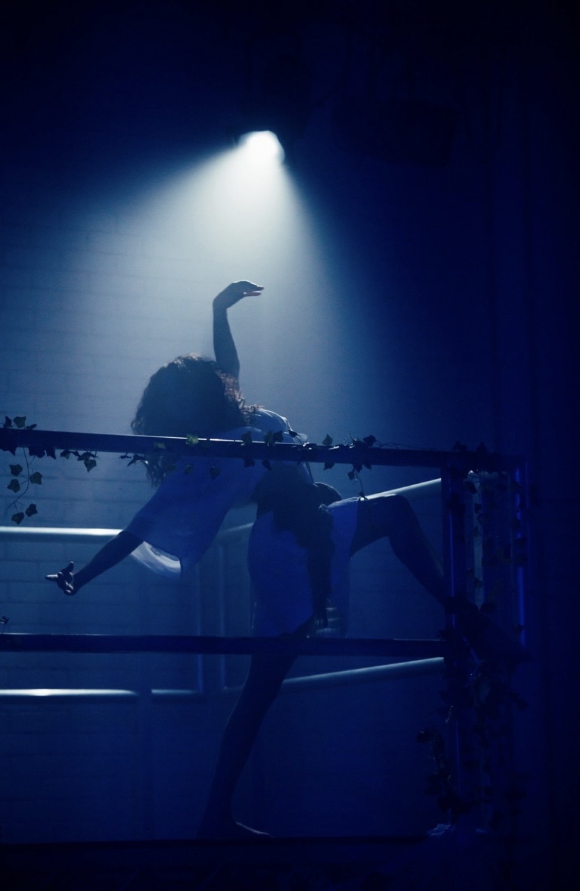 Dancer silhouette behind metal railings with ivy under a dramatic white-blue spotlight.