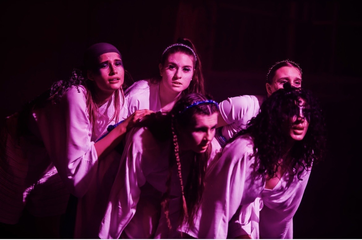 Dancers wearing white gowns under pink lighting, dark background.