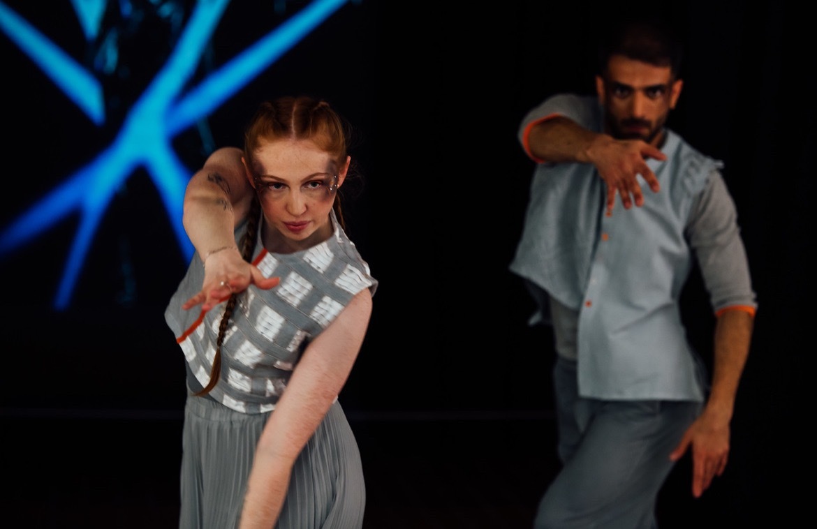 Two dancers, one in front of the other, lunging their arms forward. Dark background with blue lighting.
