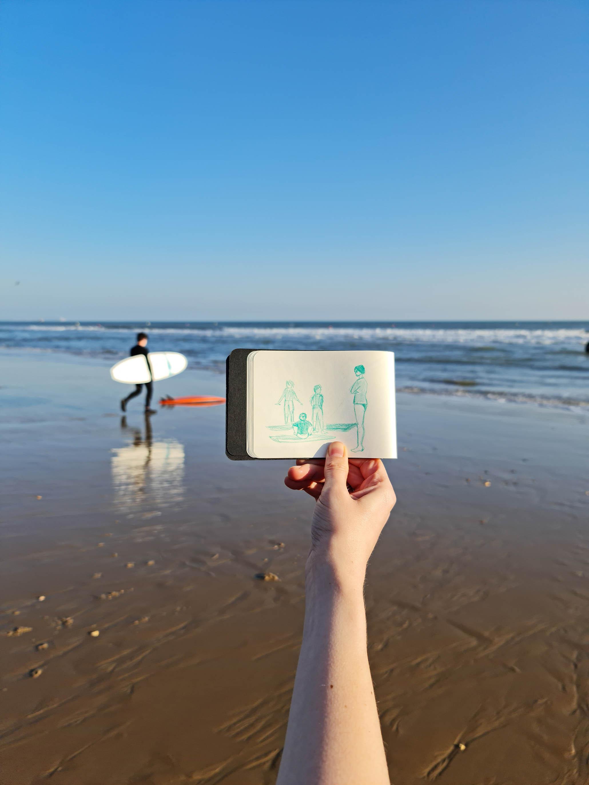 A hand at the beach holding up a small note book with an illustration of a family at the beach