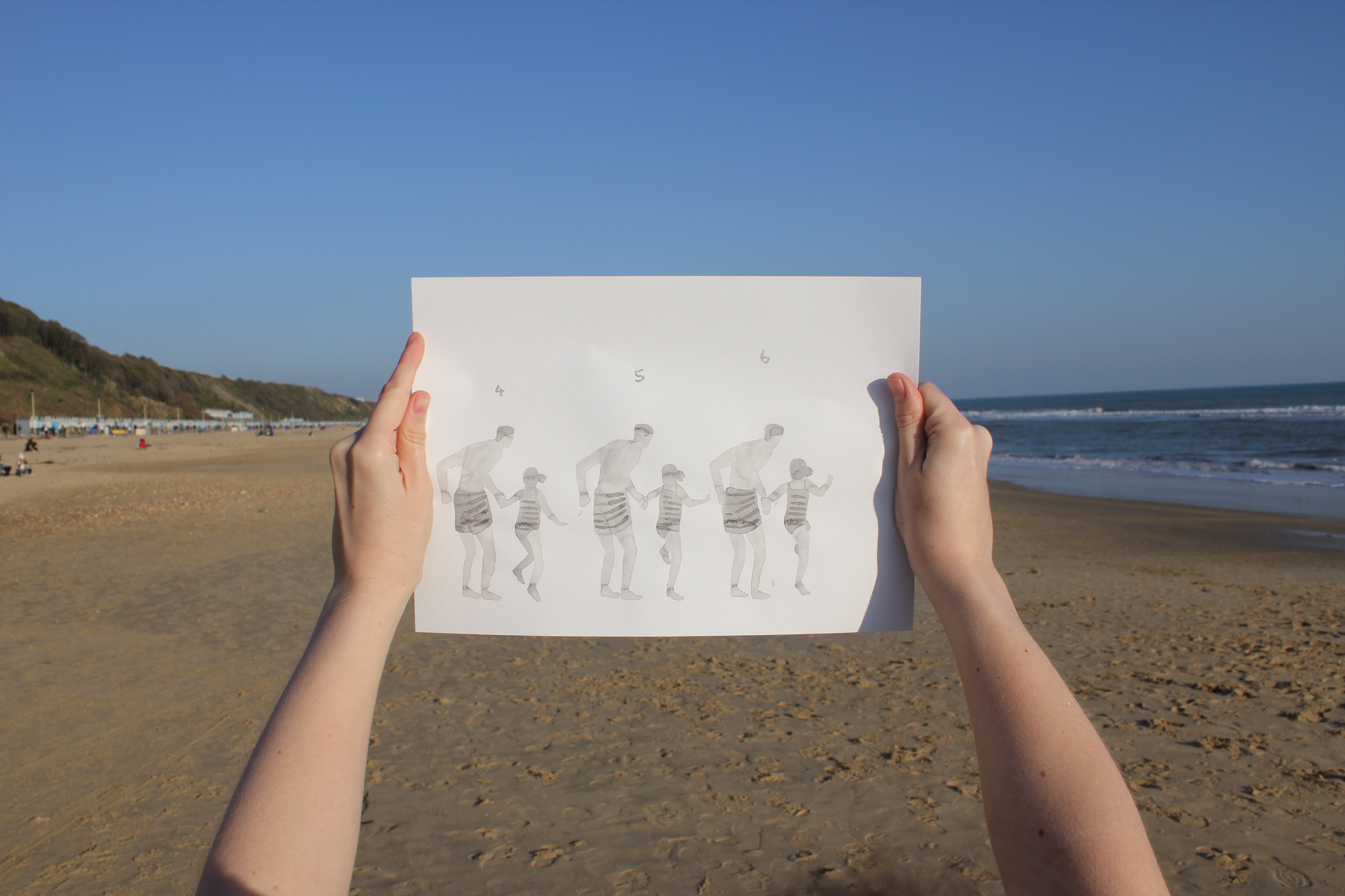 Hands at the beach holding up a small note book with an illustration of a family at the beach