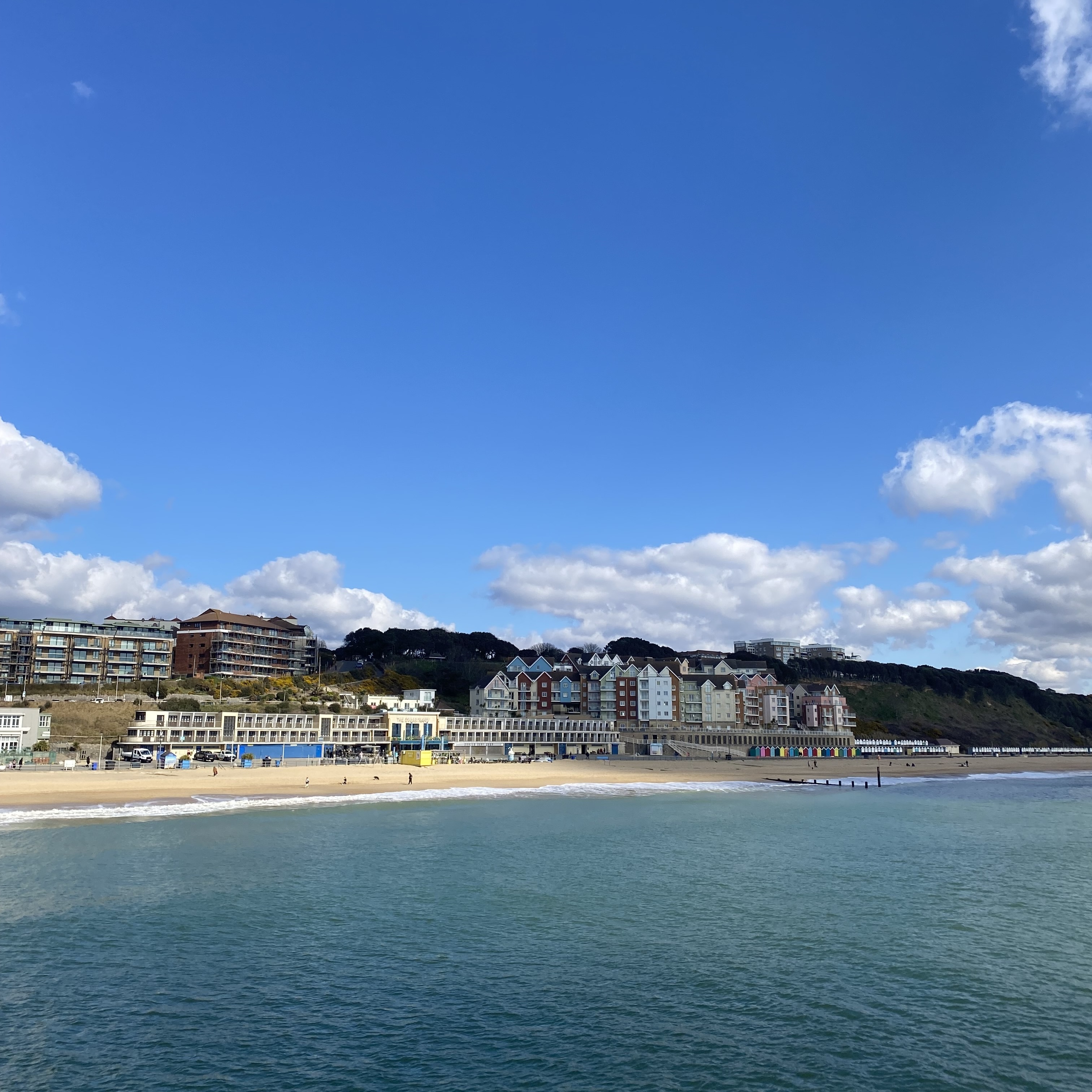 Coastal town with sandy beach, blue-green sea, and multi-storey buildings along waterfront under bright blue sky with white clouds.