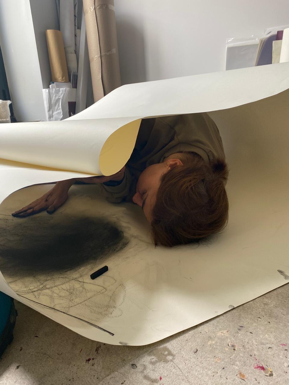 MA Fine Art graduate lying loosely wrapped in a long piece of paper, rubbing part of the paper in a circle with charcoal.