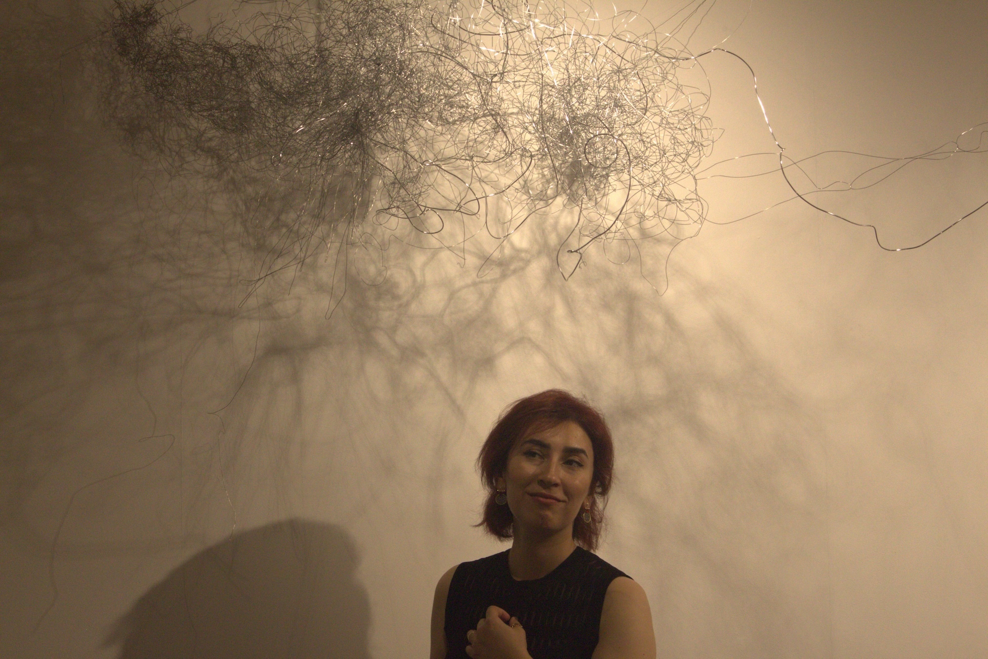 MA Fine Art student L Ghavemi standing underneath an abstract wire artwork hanging from a gallery ceiling.