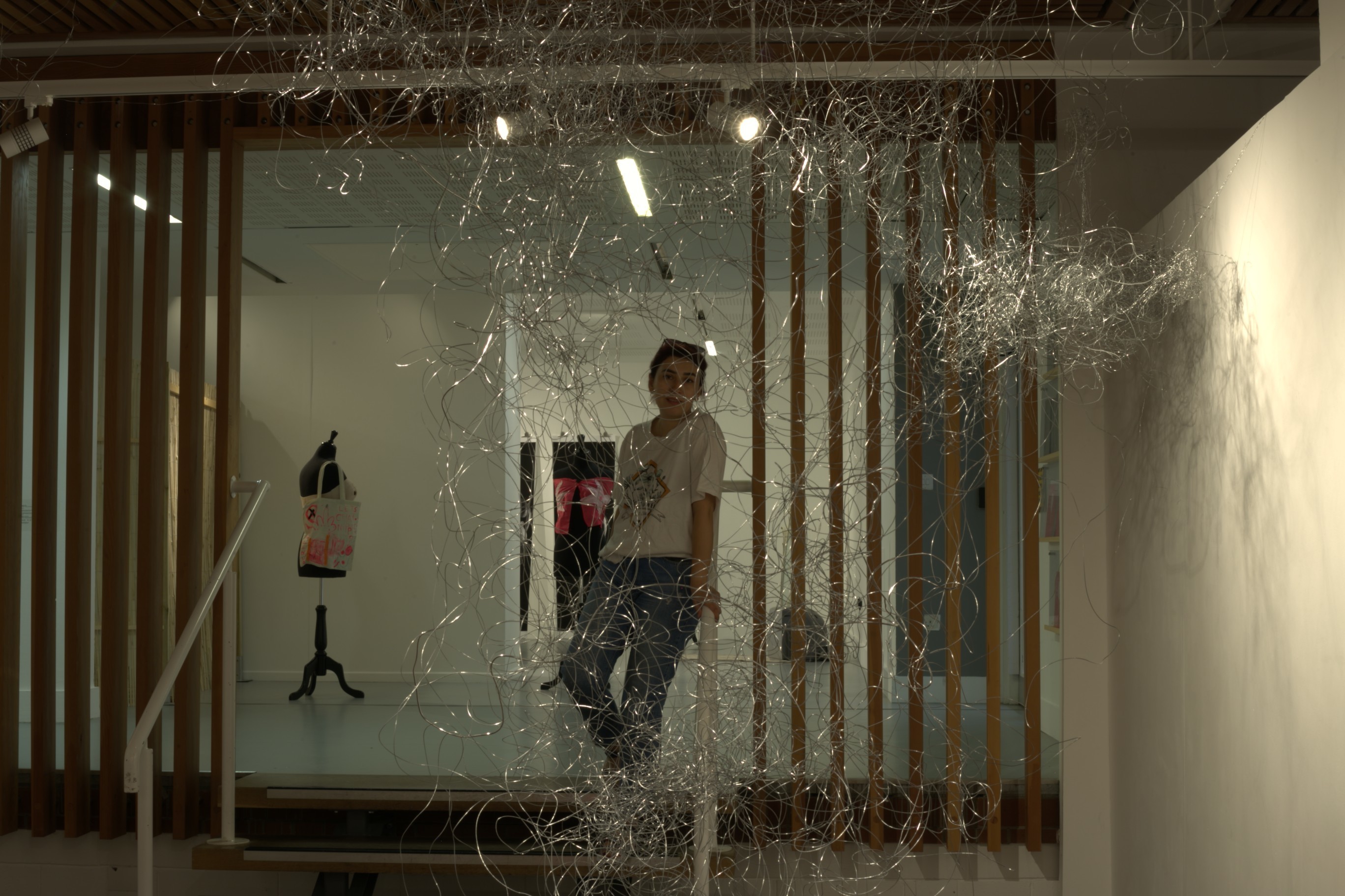 MA Fine Art graduate L Ghavemi standing on some stairs behind an abstract artwork made of a mass of wire, on display in TheGallery, Arts University Bournemouth.
