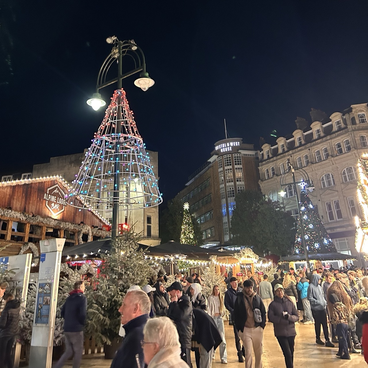 Illuminated Christmas market at night with large triangular tree structure, crowds, market stalls, and historic buildings under dark sky.