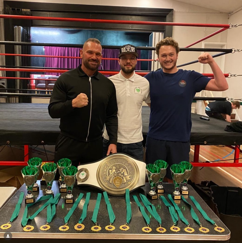 Three men posing behind boxing ring with championship belt and green ribboned medals displayed on table in front.