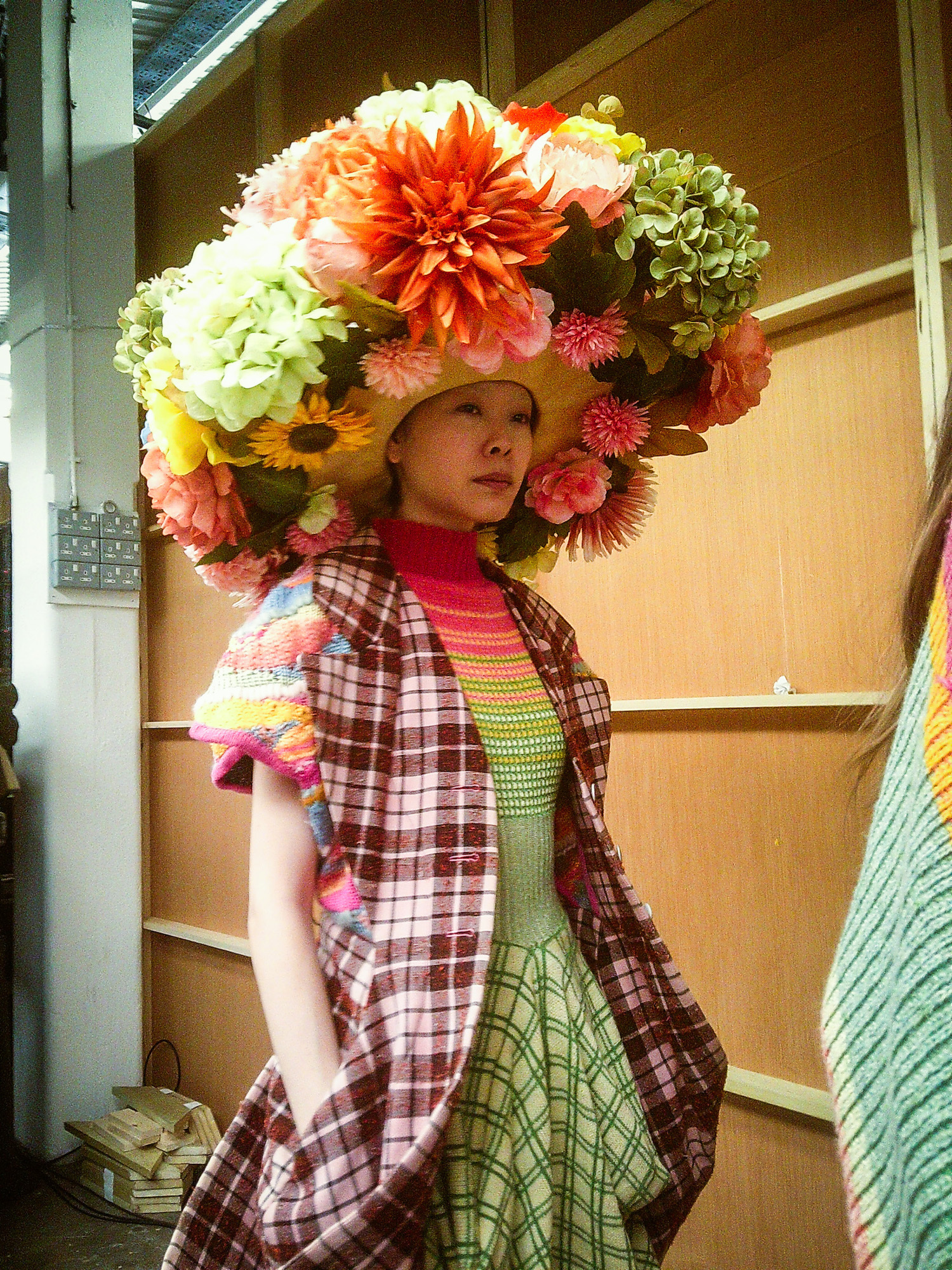 A model backstage at Graduate Fashion Week 2024 wearing colourful clothes and a large hat decorated with various flowers.