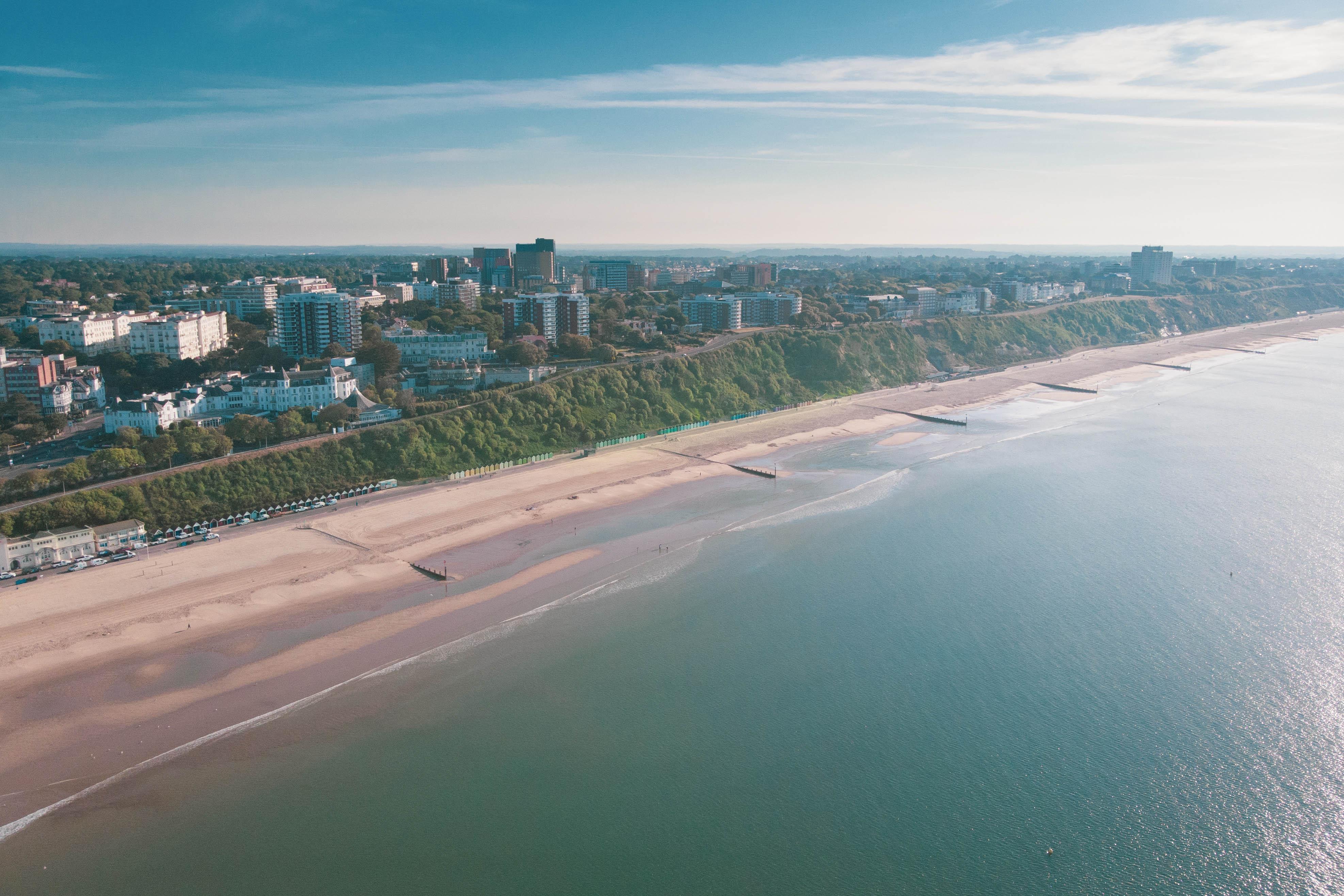 Aerial photo of a coastline with the beach, the sea, cliffs, greenery and a town further in the distance.
