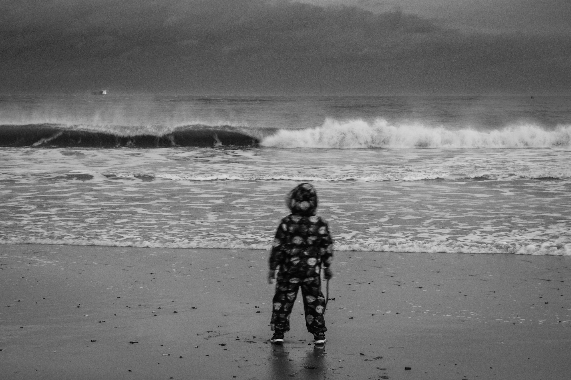 A child in patterned rainwear stands on a wet beach facing breaking waves under a cloudy sky.