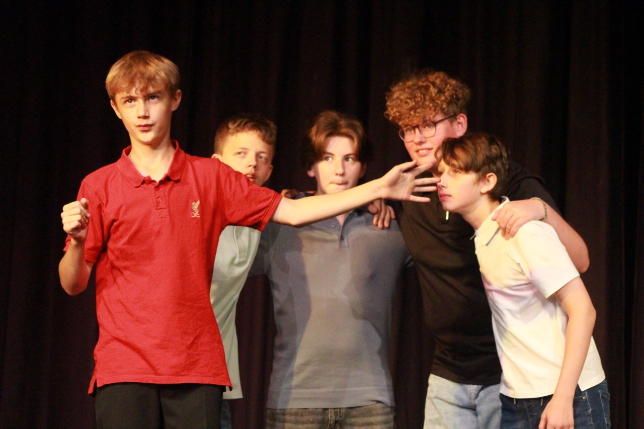 Five boys under stage lights holding onto each other, student on the left reaching out in a red top.