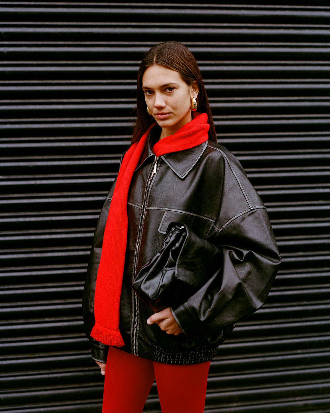 Woman in oversized black leather jacket and bright red scarf holding black handbag against corrugated metal shutters.