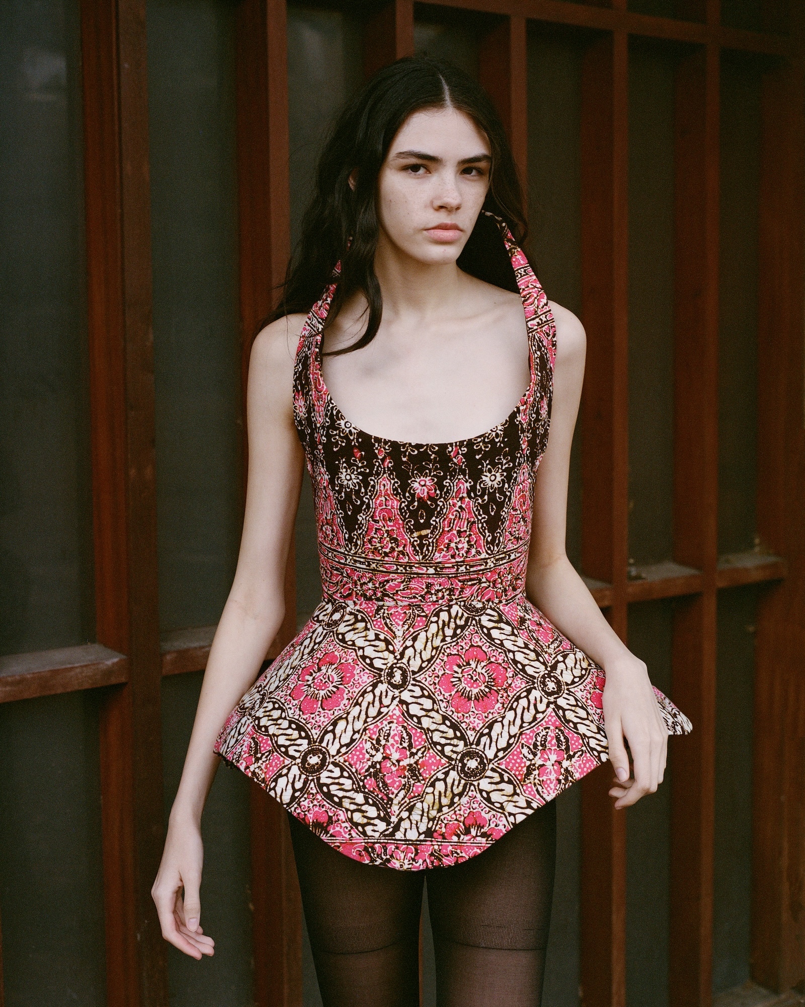 Woman with dark hair wearing patterned pink and brown sleeveless top standing in front of wooden glass doors.