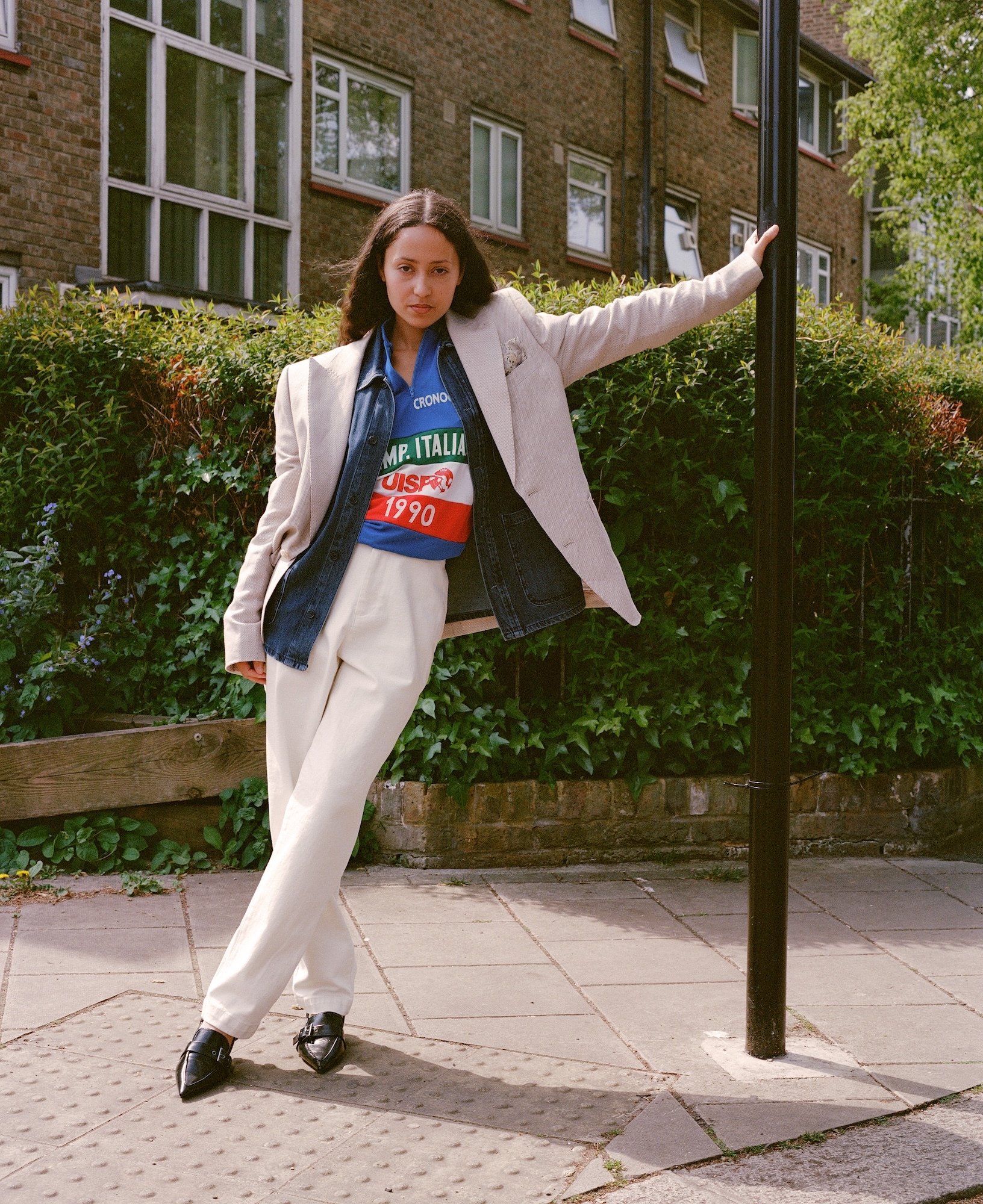 Woman in beige suit and blue graphic t-shirt posing against lamp post on pavement, brick buildings with ivy in background.