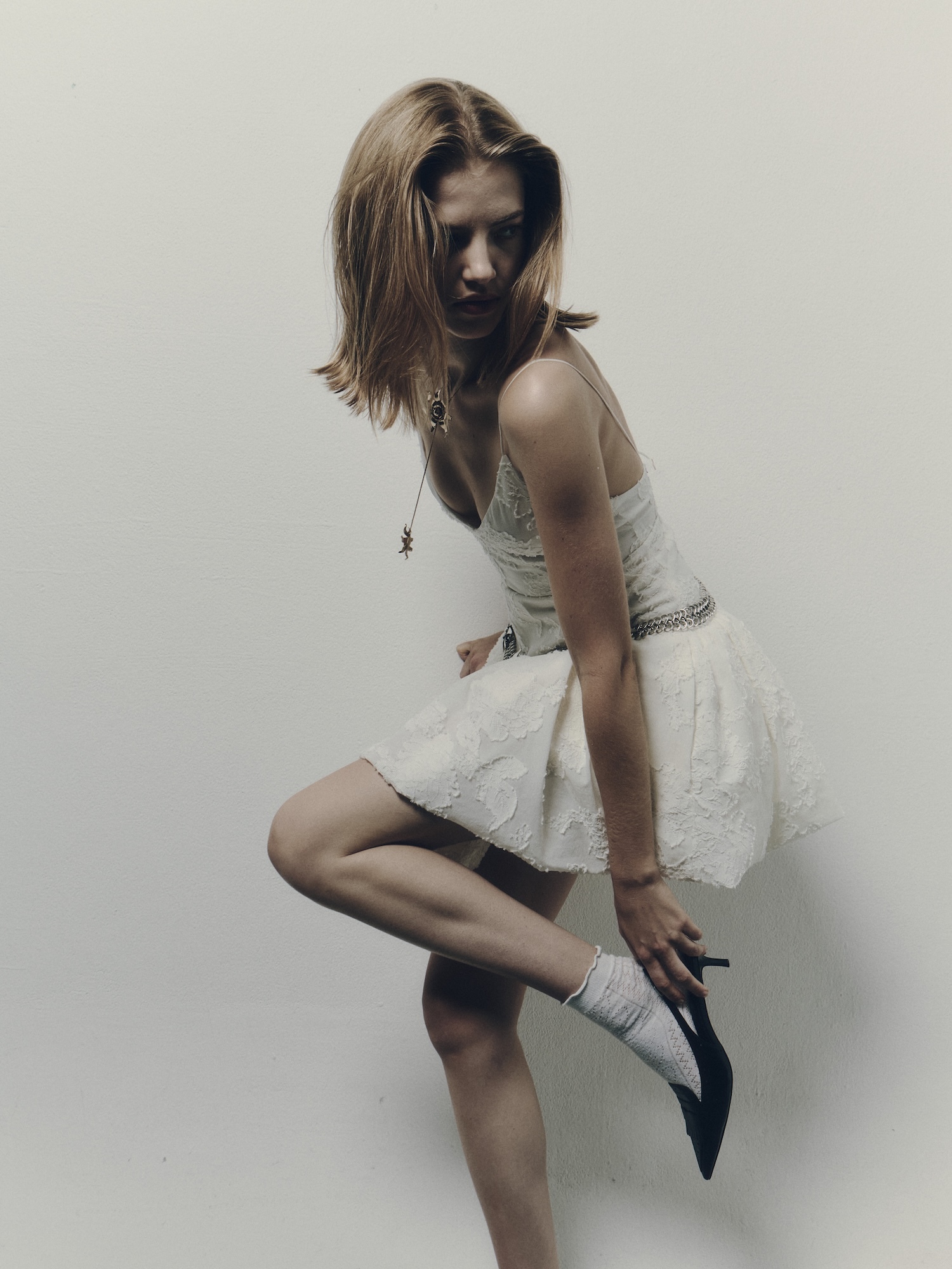 Woman with blonde bob hairstyle wearing white dress and black shoes, bending forward against neutral background.