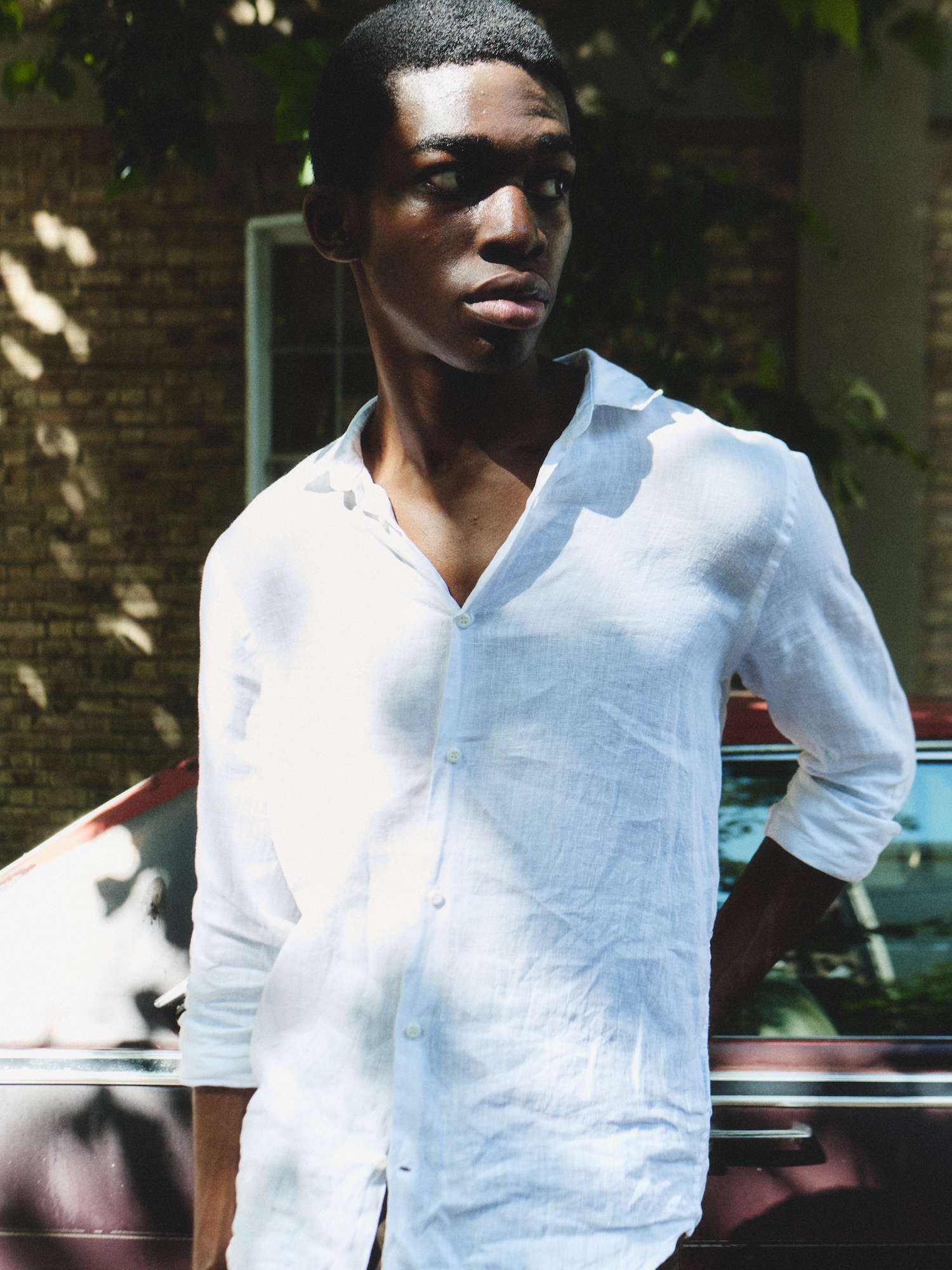 Young Black man in white long-sleeved shirt outdoors with dappled sunlight and shadows on his face, blurred background.