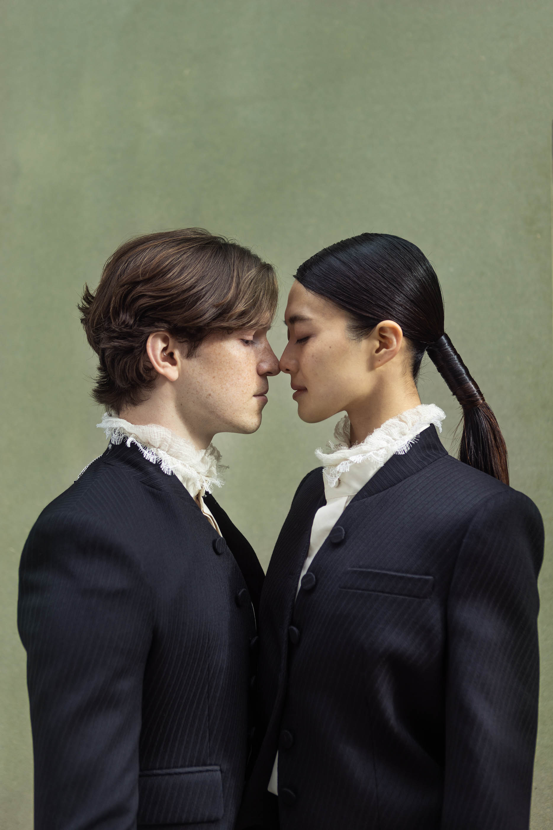 A man and woman in matching dark tailored suits face each other with their noses touching on a plain green background.