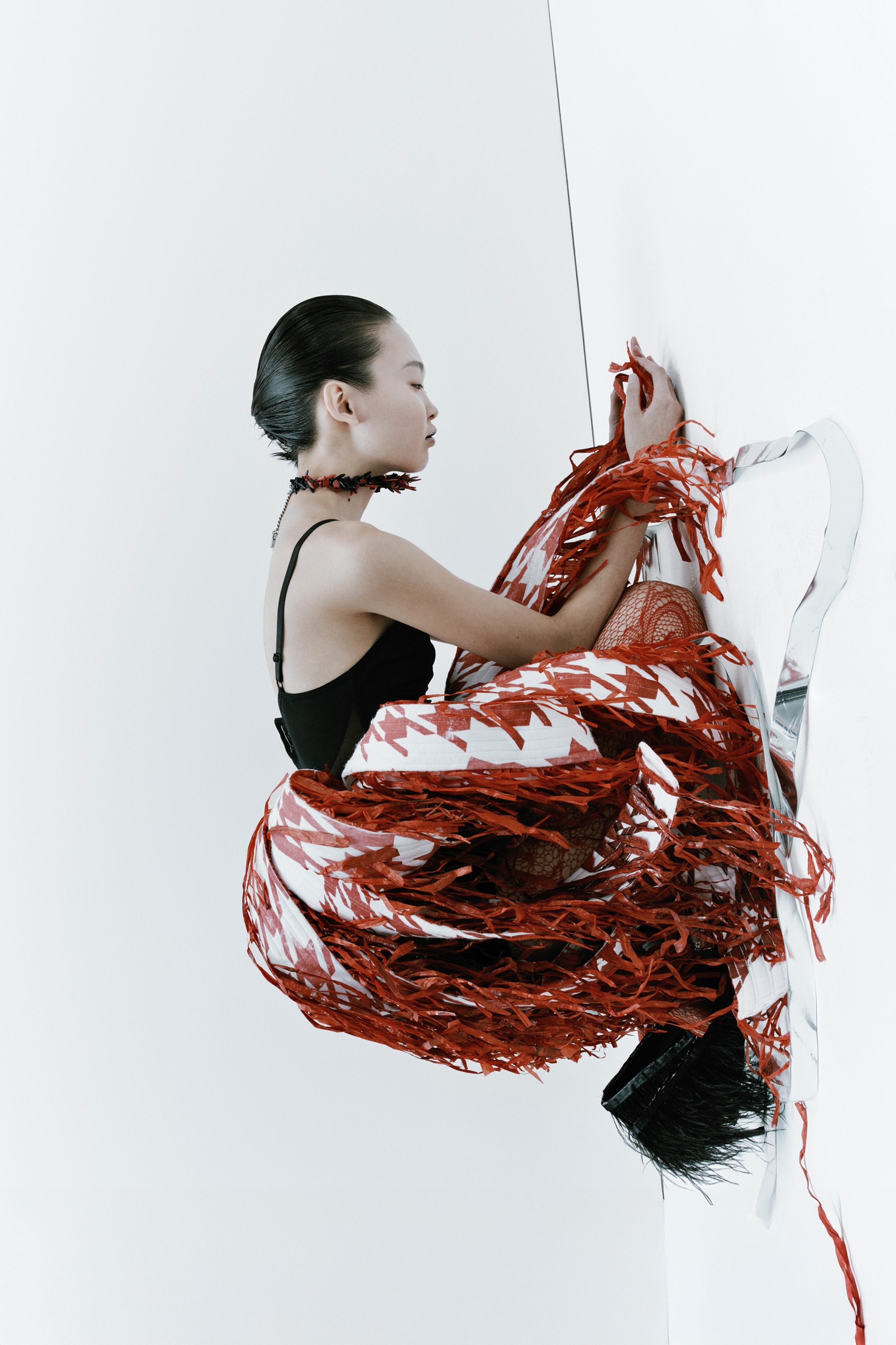 A woman in a black leotard is suspended against a white wall with flowing red-and-white fabric wrapped around her body.