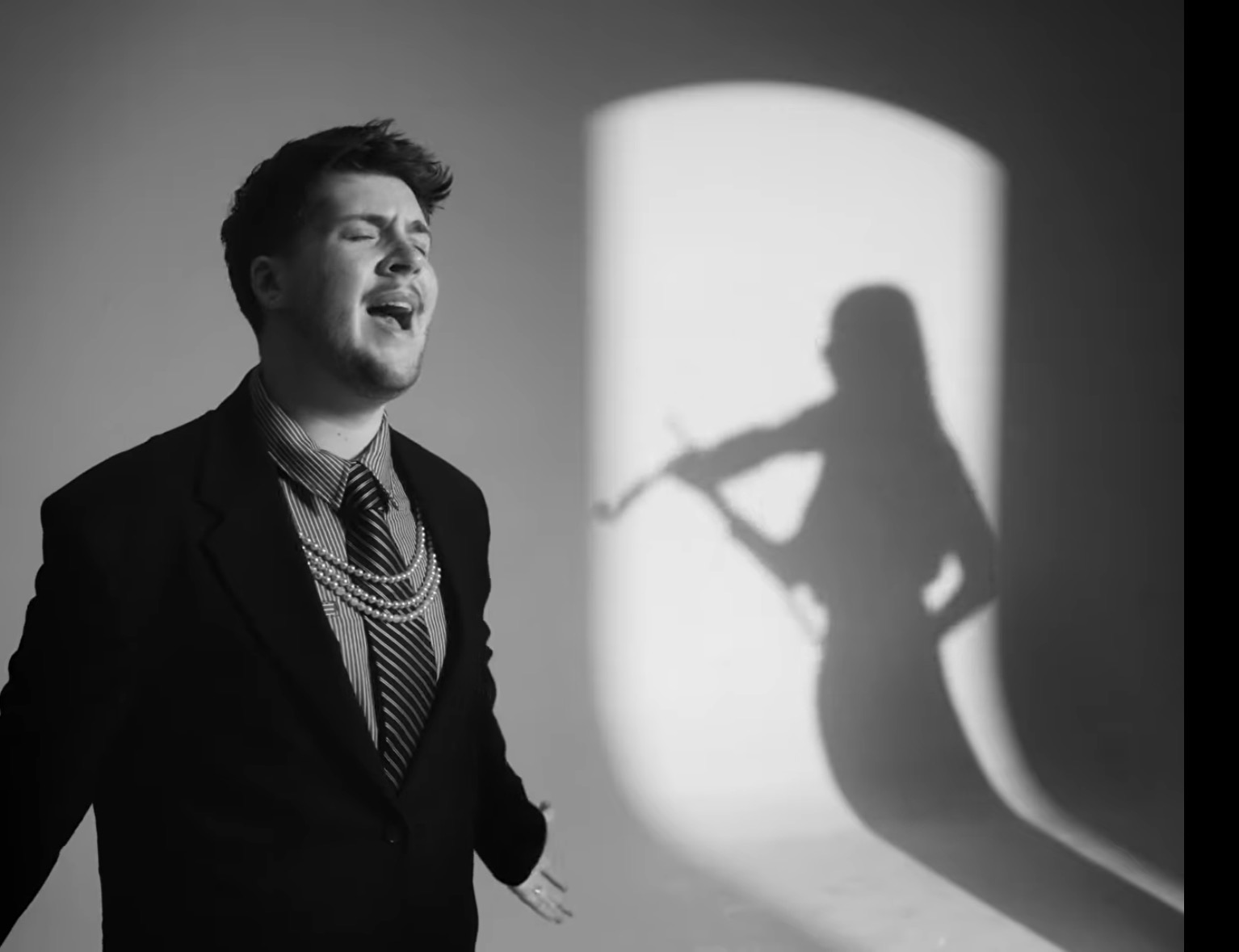 A man in a suit and striped tie singing beside a bright backlit panel, with his shadow cast as a violinist on the wall.