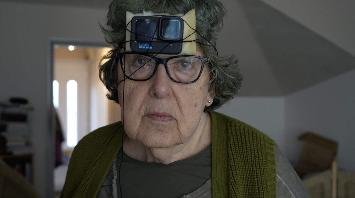 An elderly woman with glasses and grey hair is shown with a camera mounted on her forehead. She is wearing a green cardigan over a grey top, and the background features blurred domestic items and light from windows.