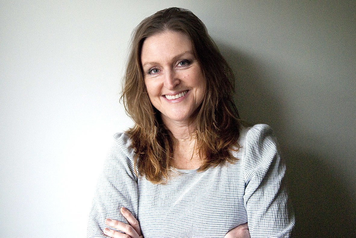 Woman with brown hair smiling, arms crossed, wearing grey and white striped top against white wall.