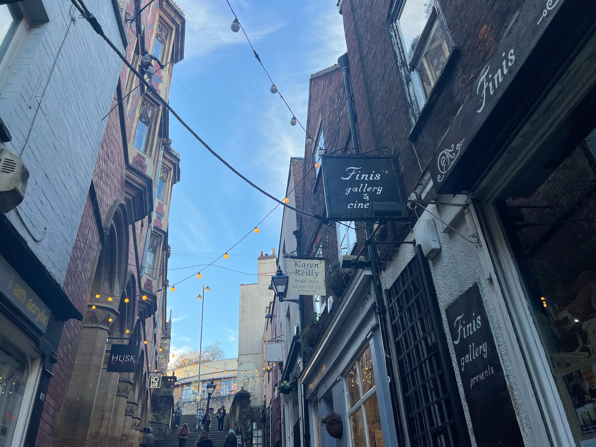Shop fronts and signs on either side of a Bristol high street, with a cloudy blue sky above.