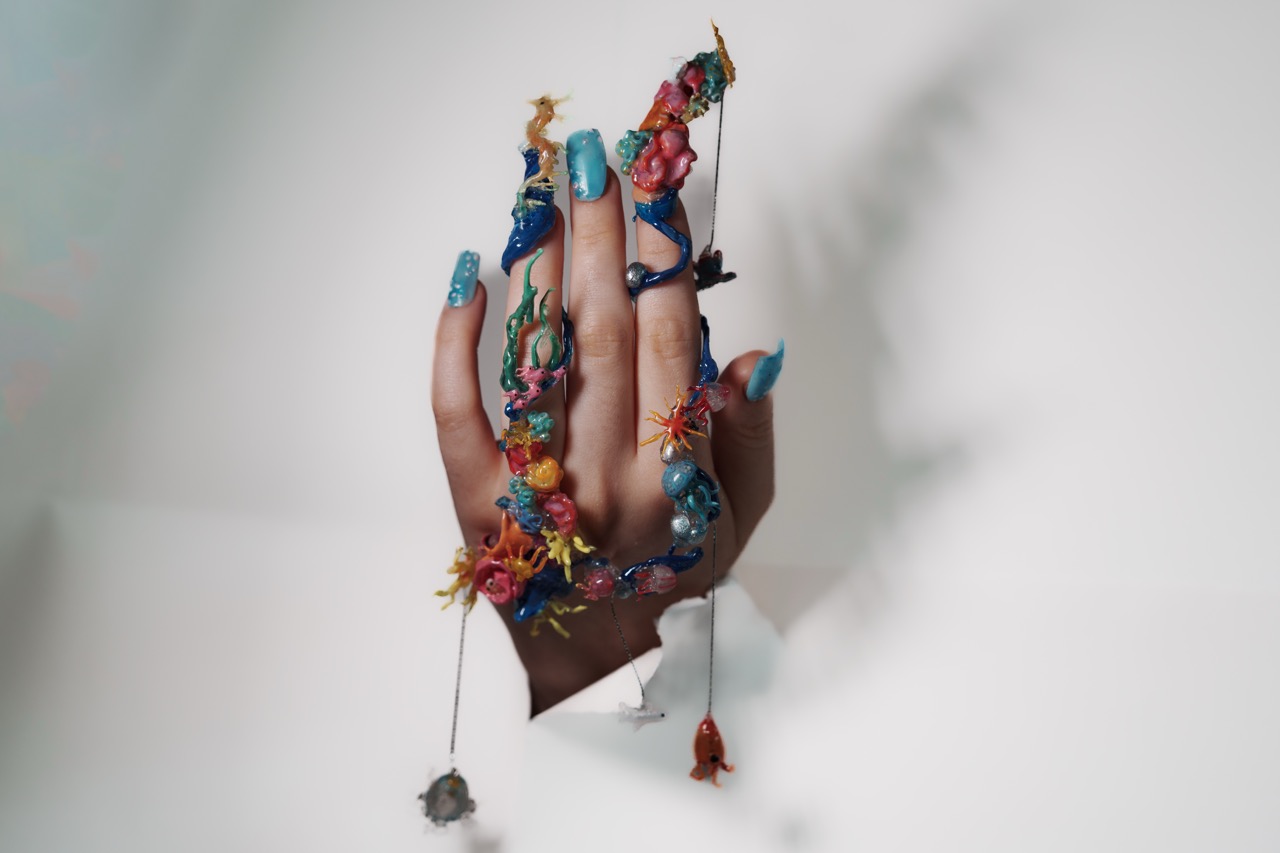 A hand with blue-painted nails emerges through a torn white surface, decorated with colourful flowers and ornaments.
