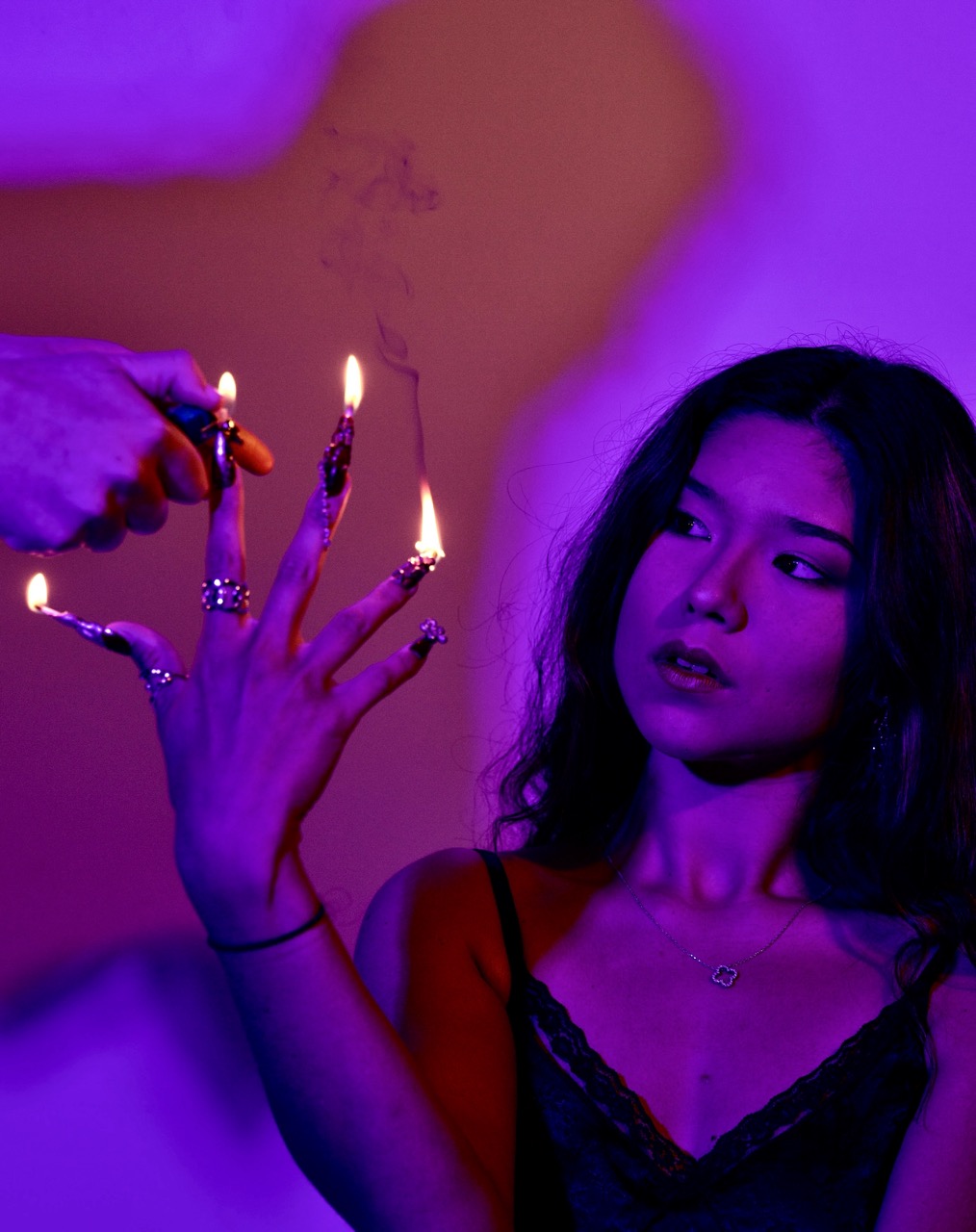 A woman in a black lace top holds up her hand with lit cigarette lighter flames on her fingers against a purple background.
