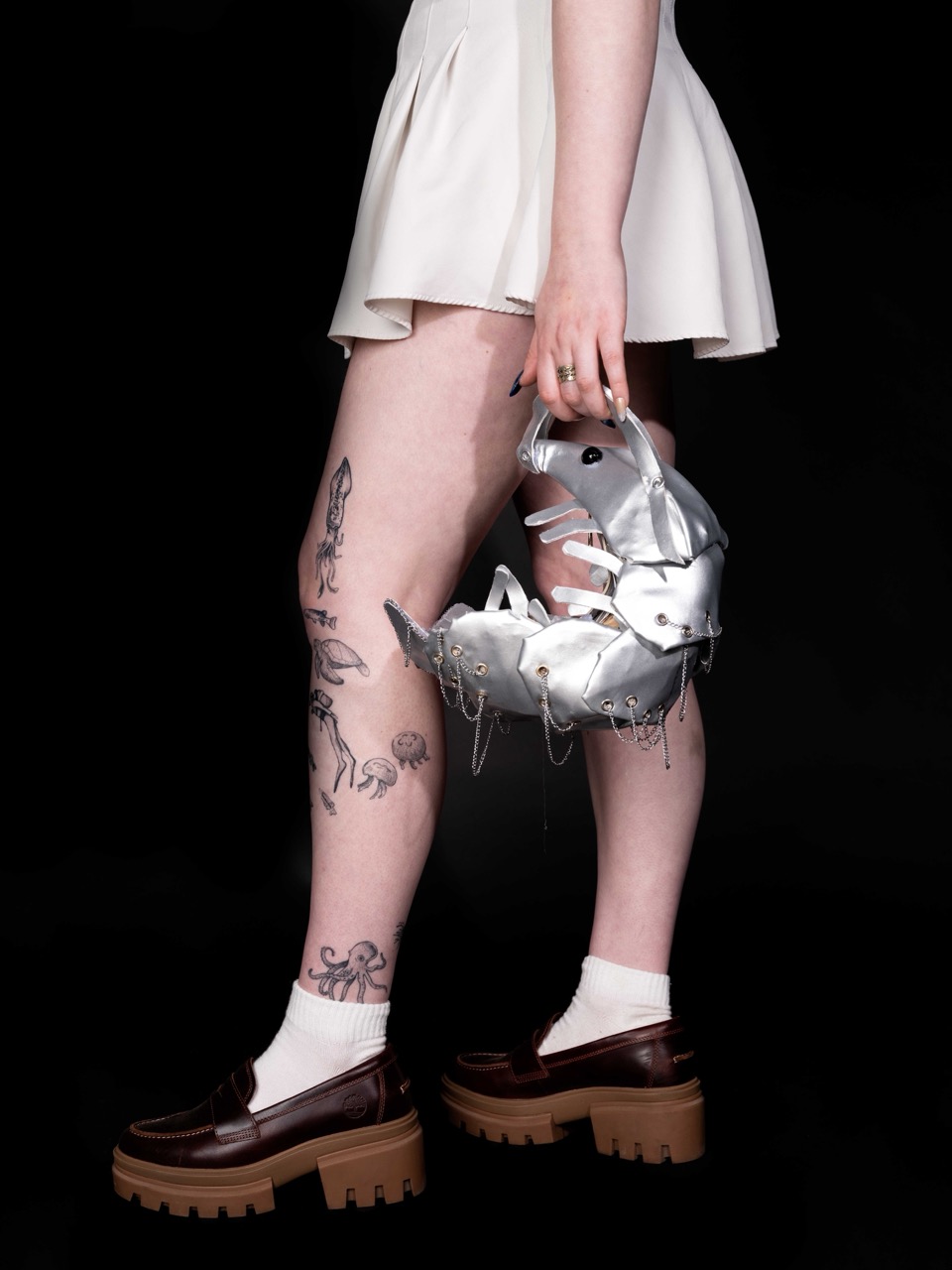 A woman in a white skirt and socks holds a metallic sculptural handbag beside tattooed legs, wearing brown chunky loafers against a black background.