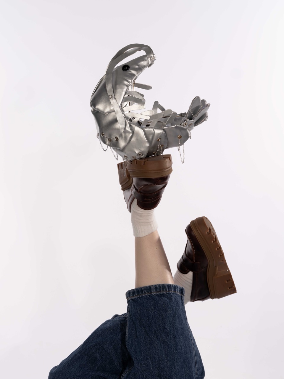 A person’s raised leg wearing blue jeans and a brown shoe holds a metallic silver robotic foot, against a plain white background.