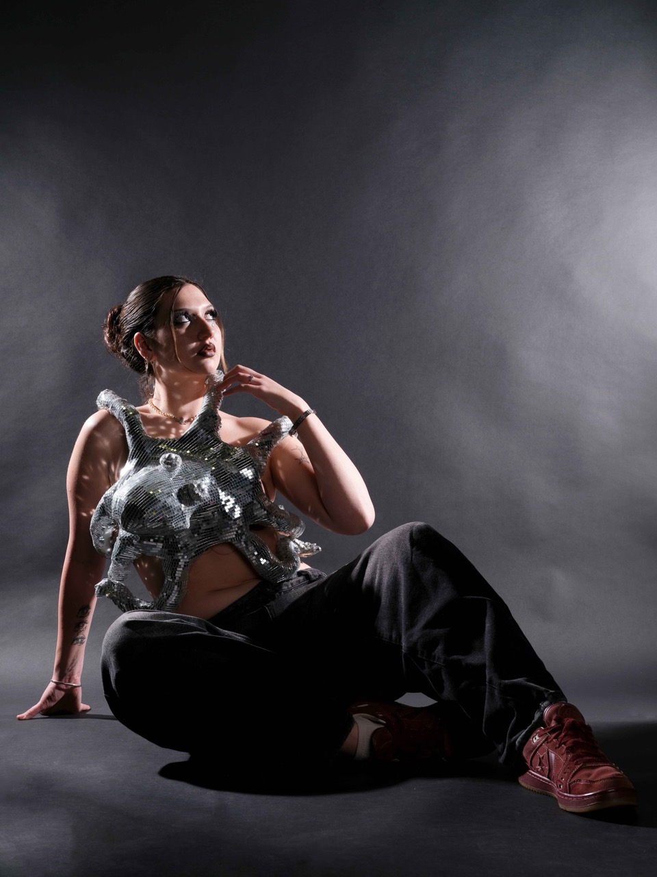 A seated woman wearing a reflective silver top and black trousers poses against a dark studio backdrop with dramatic side lighting.