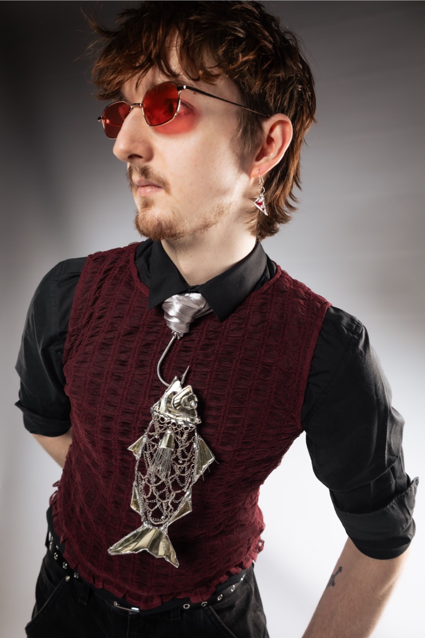 A man wearing red-tinted glasses, a burgundy vest and a black shirt stands against a plain grey background, with a silver fish-shaped pendant on a chain.