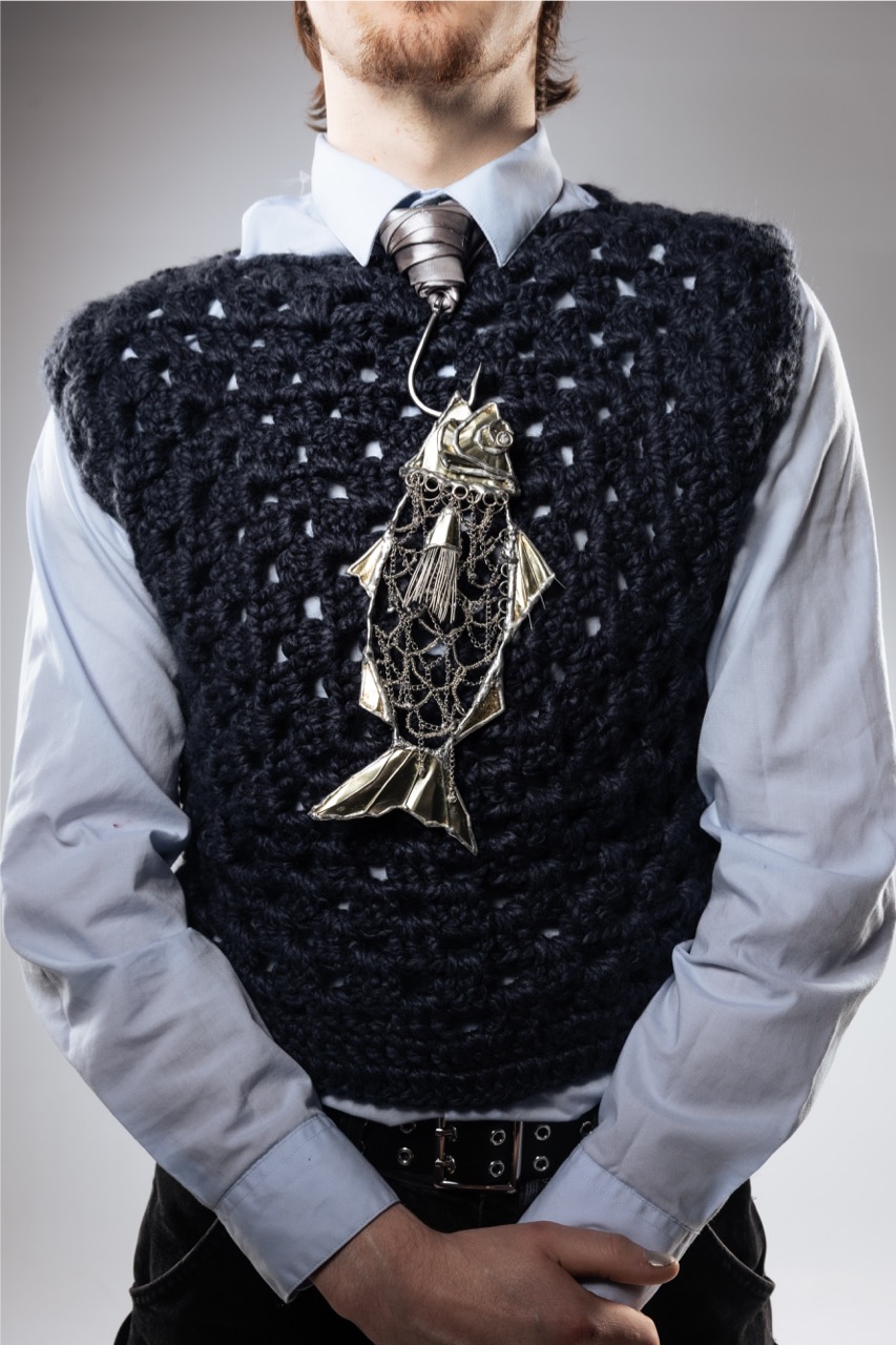 A man wearing a light blue shirt, striped tie and black knitted vest with a fish-shaped ornament, photographed against a plain grey background.