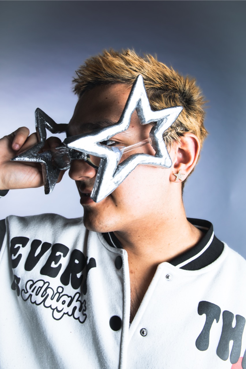 A young man with bleached hair wears oversized star-shaped novelty glasses and a white varsity jacket against a plain grey background.